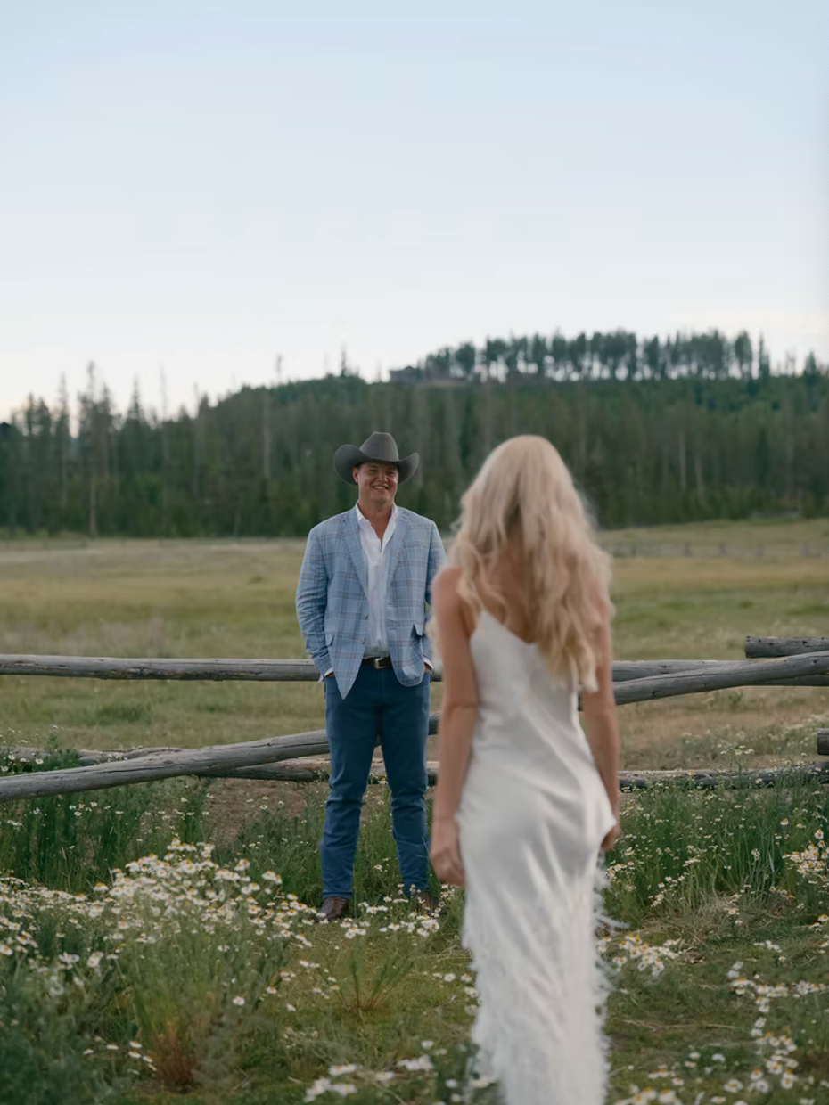 A couple in western attire stands near a wooden fence in a meadow filled with wildflowers during sunset.
