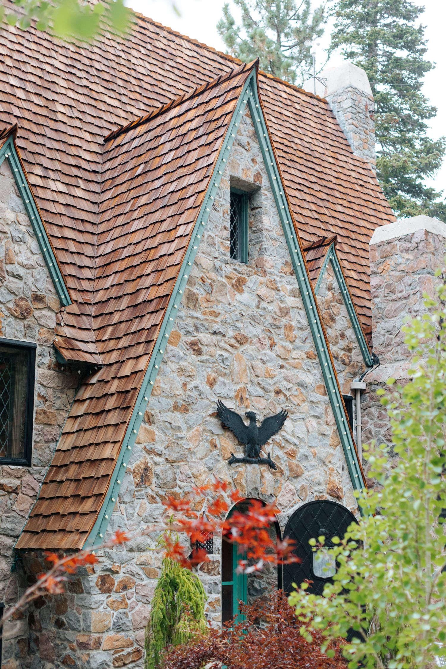 Stone cottage with steep shingled roof and green trim features vibrant red fall foliage.