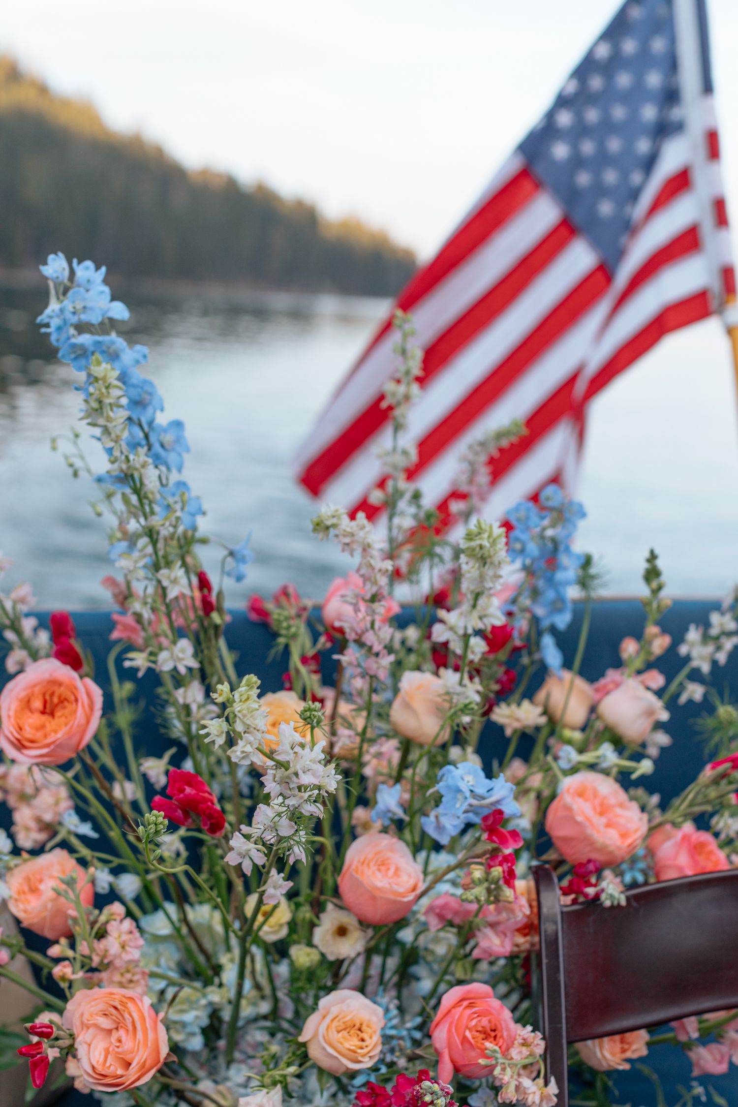 American flags wave above a romantic floral arrangement of pink roses and blue delphinium by the water.