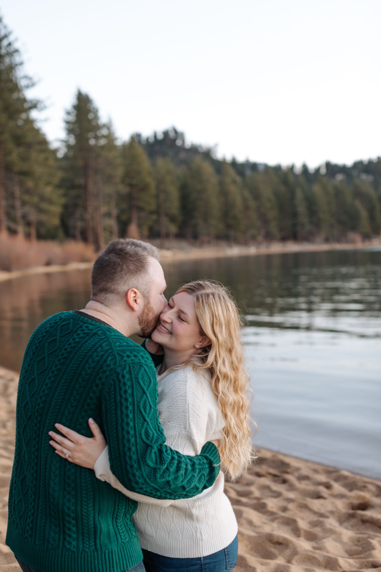 Intimate lakeside photoshoot featuring a couple in cozy winter sweaters against a mountain backdrop.