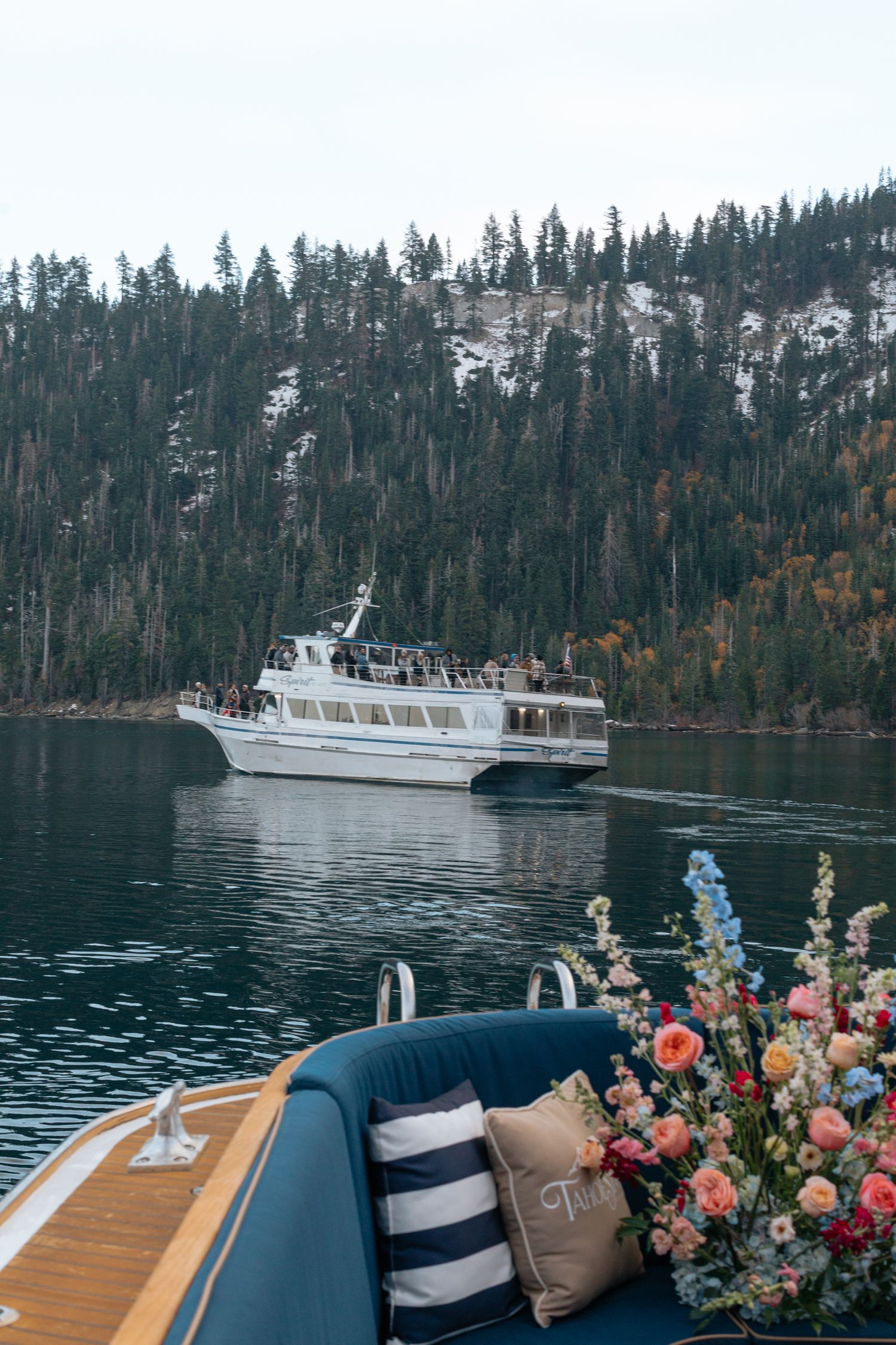 White tour boat cruising on lake surrounded by snow-dusted evergreen forest with floral decorations in foreground.