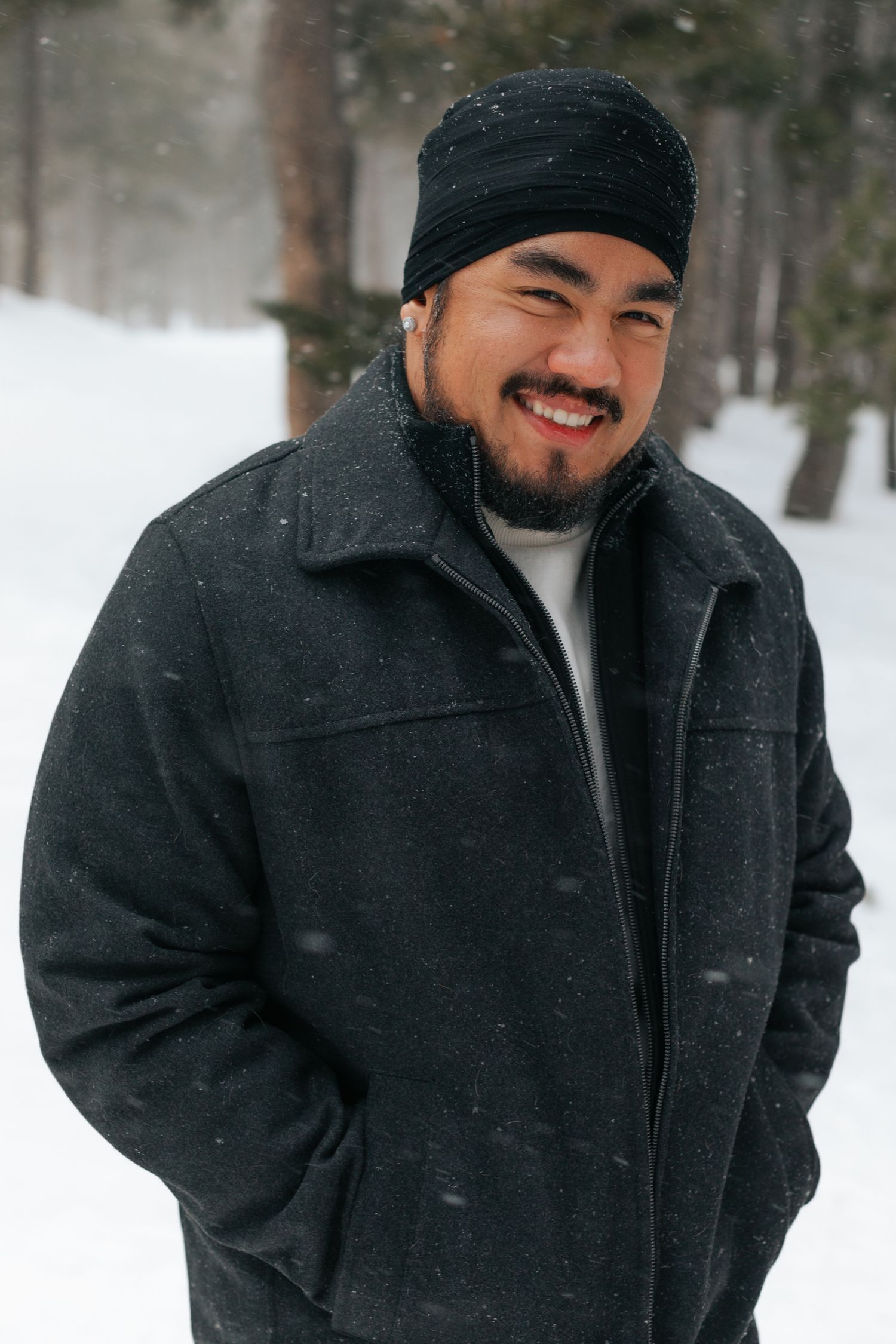 Person in black winter coat and beanie smiles warmly during a snowy outdoor portrait session.
