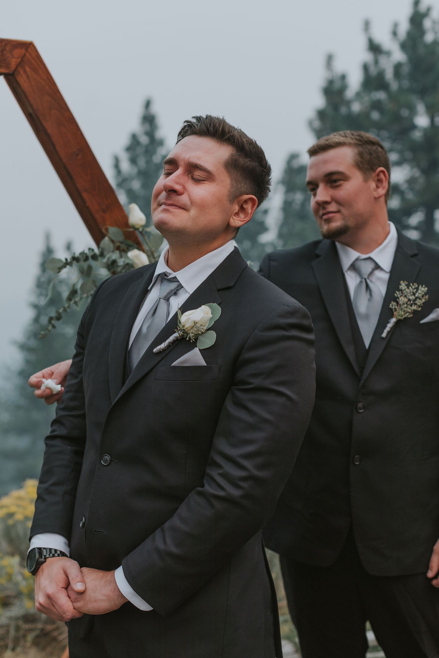Groom in dark suit with white boutonniere closes eyes emotionally at outdoor wedding ceremony, groomsman smiling behind him.