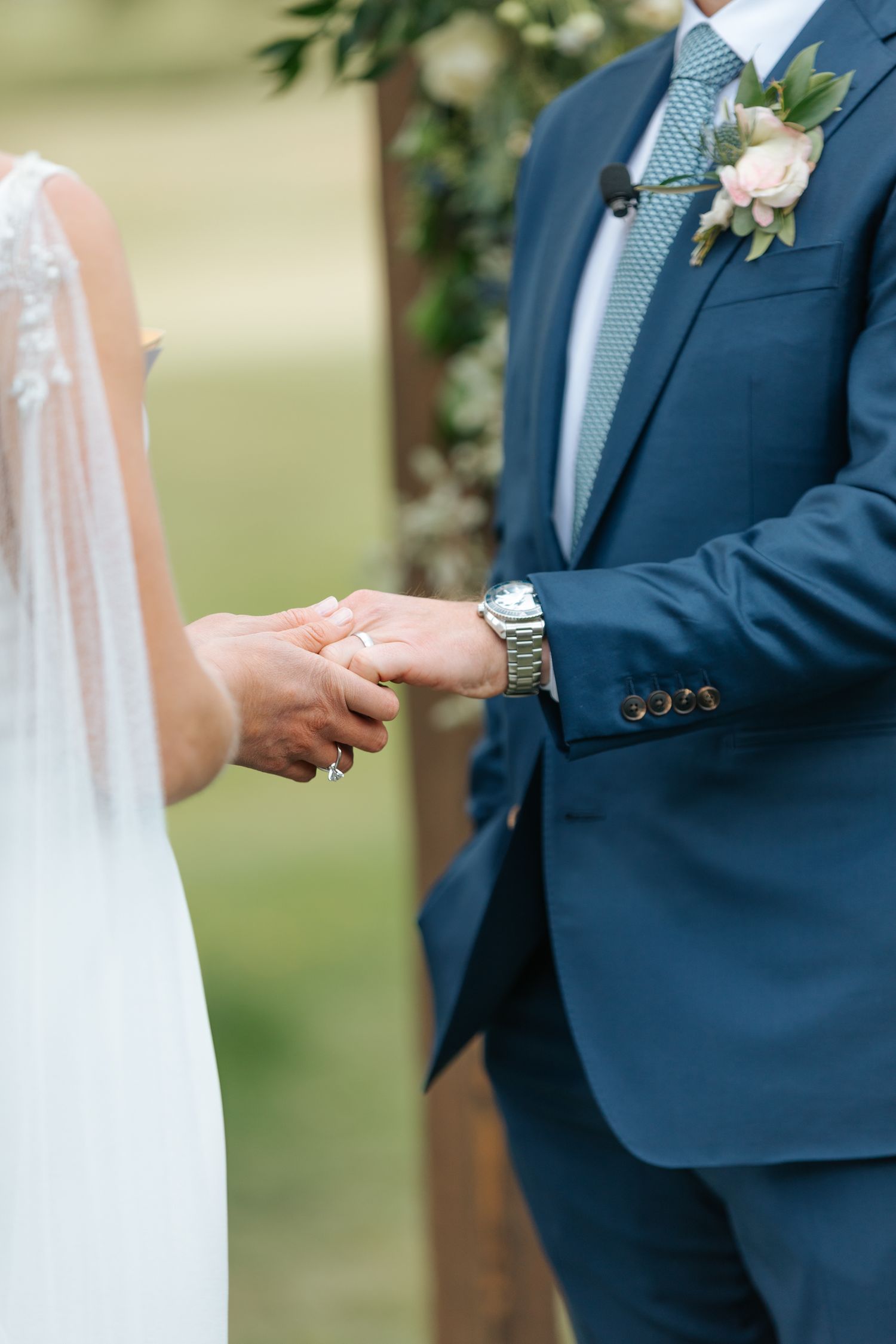 Close-up details of wedding ceremony showing hands, rings, and navy blue suits.