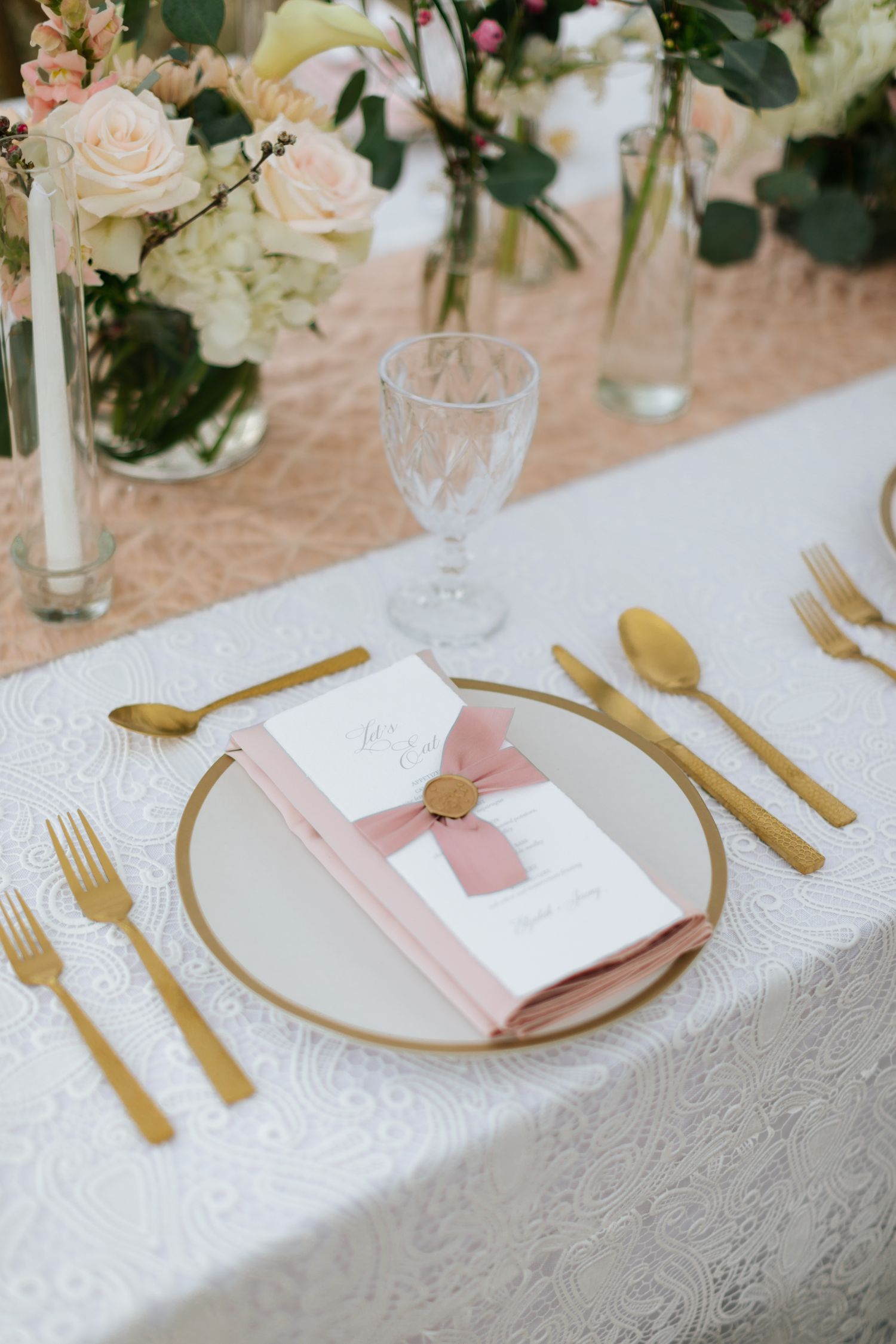 Elegant place setting with gold cutlery, white plates, and pink napkins on a white lace tablecloth.