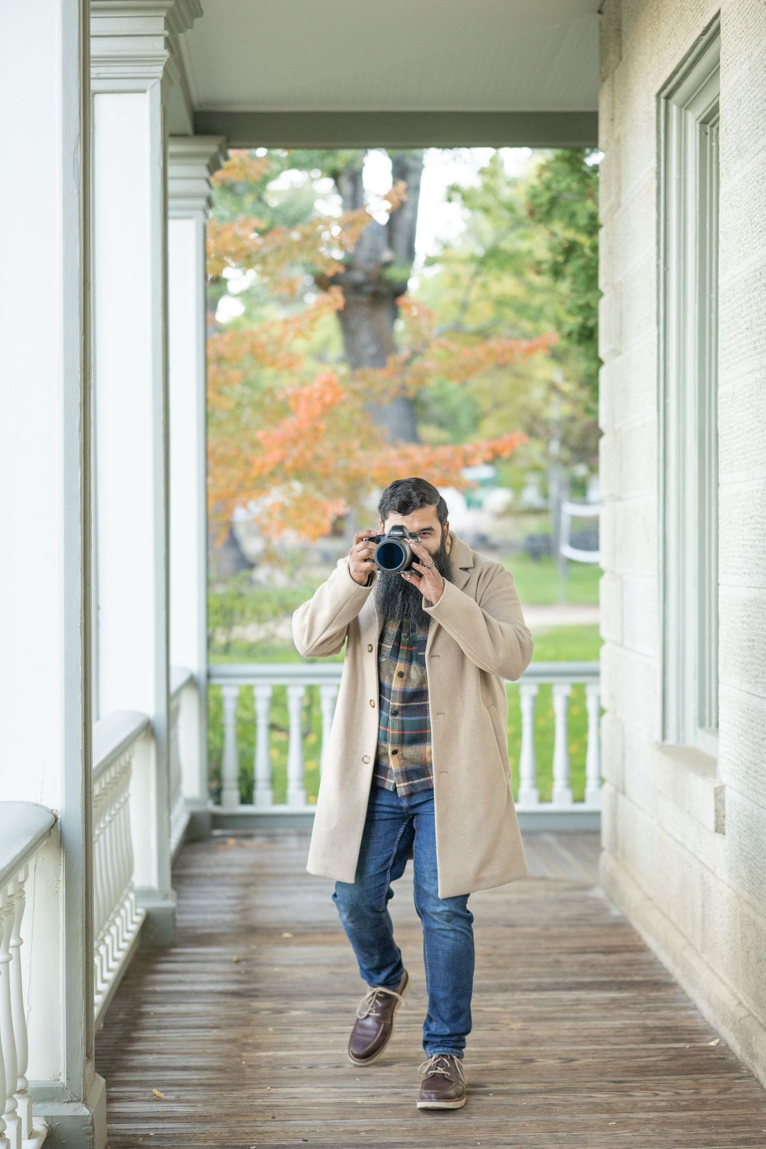 Bearded man in beige coat photographs with DSLR camera on wooden porch surrounded by autumn foliage.