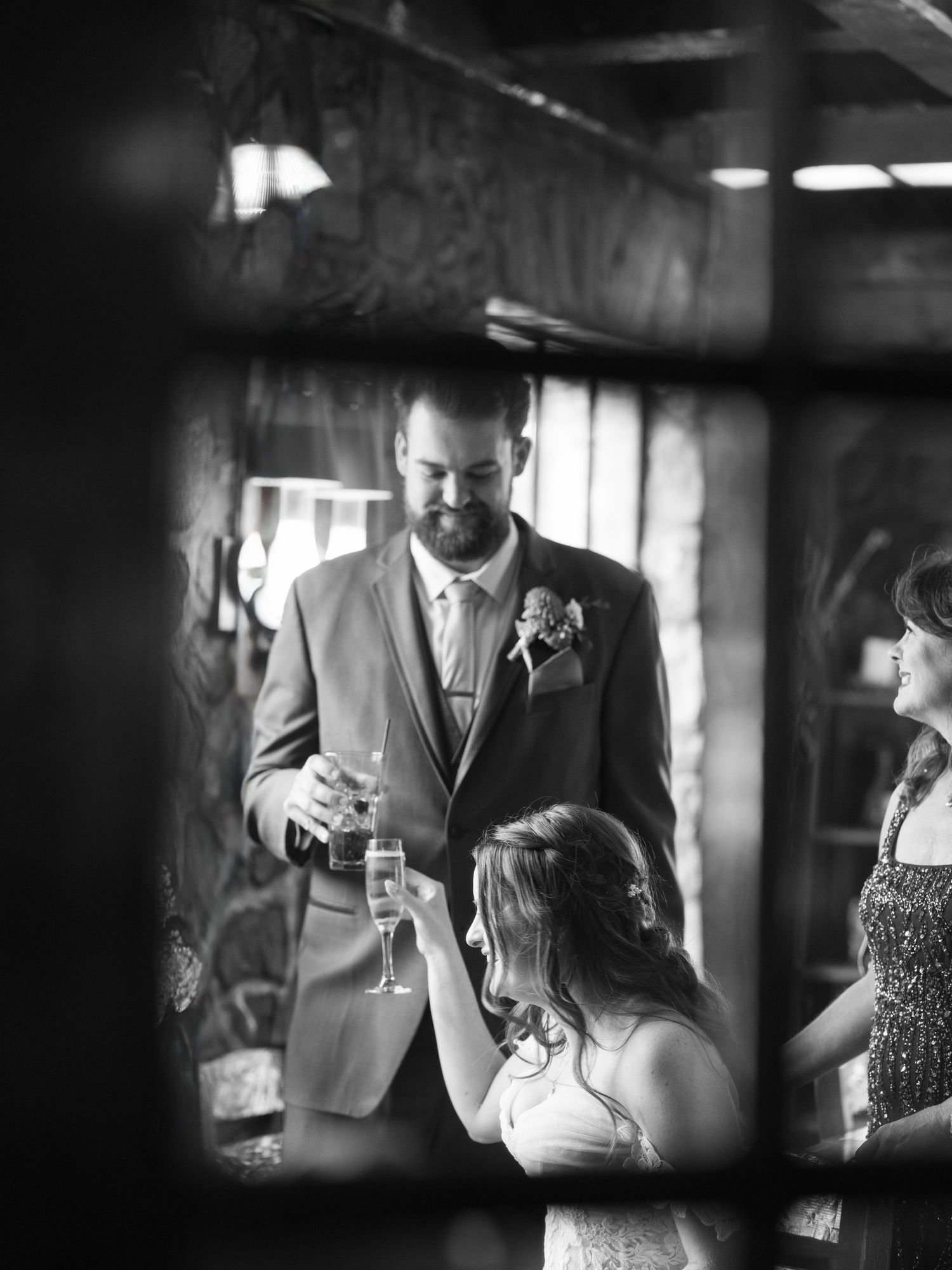 Black and white wedding photo of groom in suit with boutonniere toasting with bride in elegant venue.