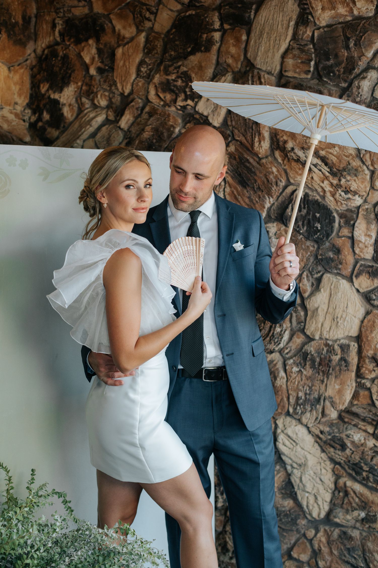 Couple sharing intimate moment under white umbrella against stone wall backdrop.