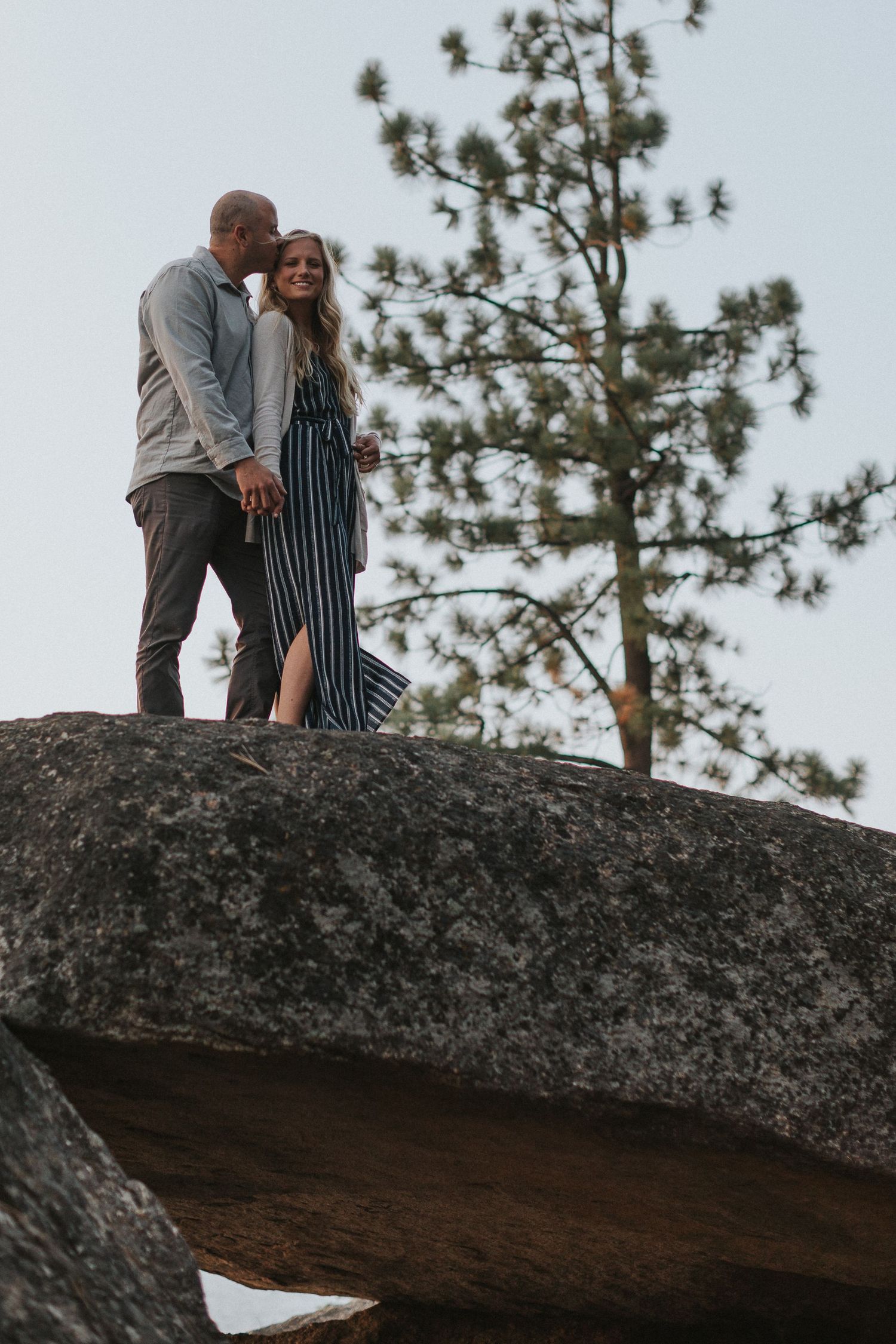 Couple sharing a romantic moment on a large rock formation with pine trees in the background.