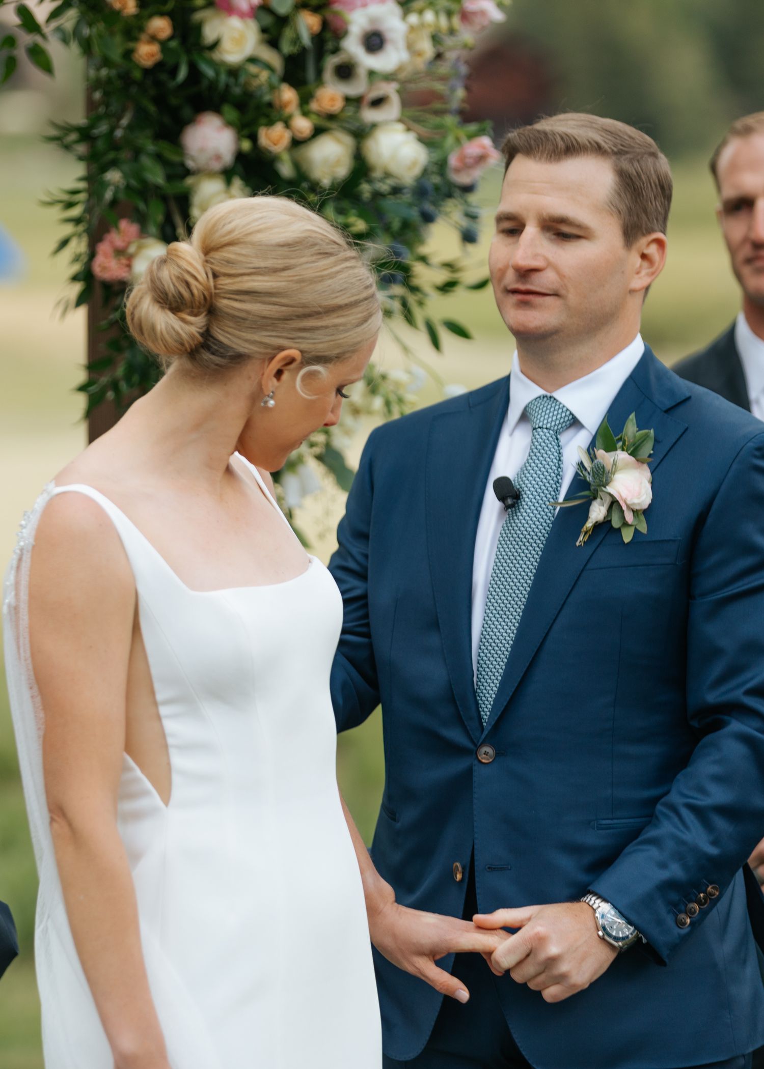 Bride and groom exchange vows outdoors at elegant wedding ceremony, groom in navy suit with floral boutonniere.