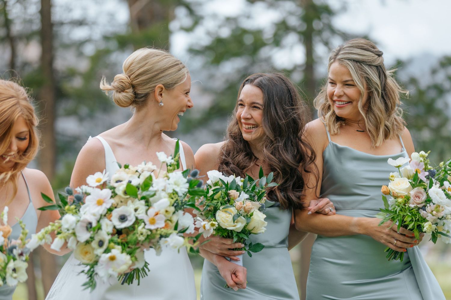 Bridesmaids in sage green dresses hold white and blush floral bouquets at an outdoor wedding ceremony.