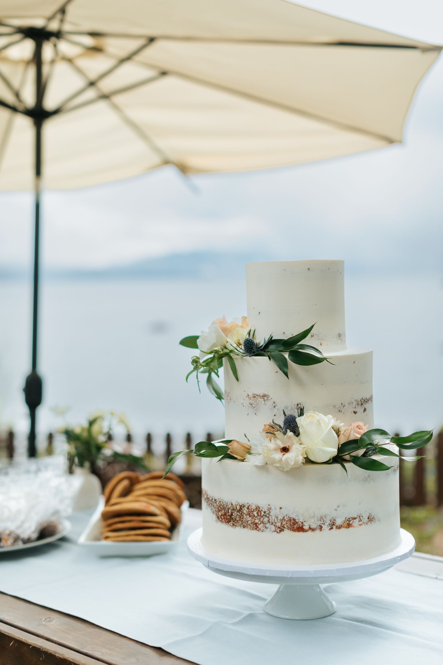 White wedding cake decorated with fresh flowers displayed under outdoor umbrellas.