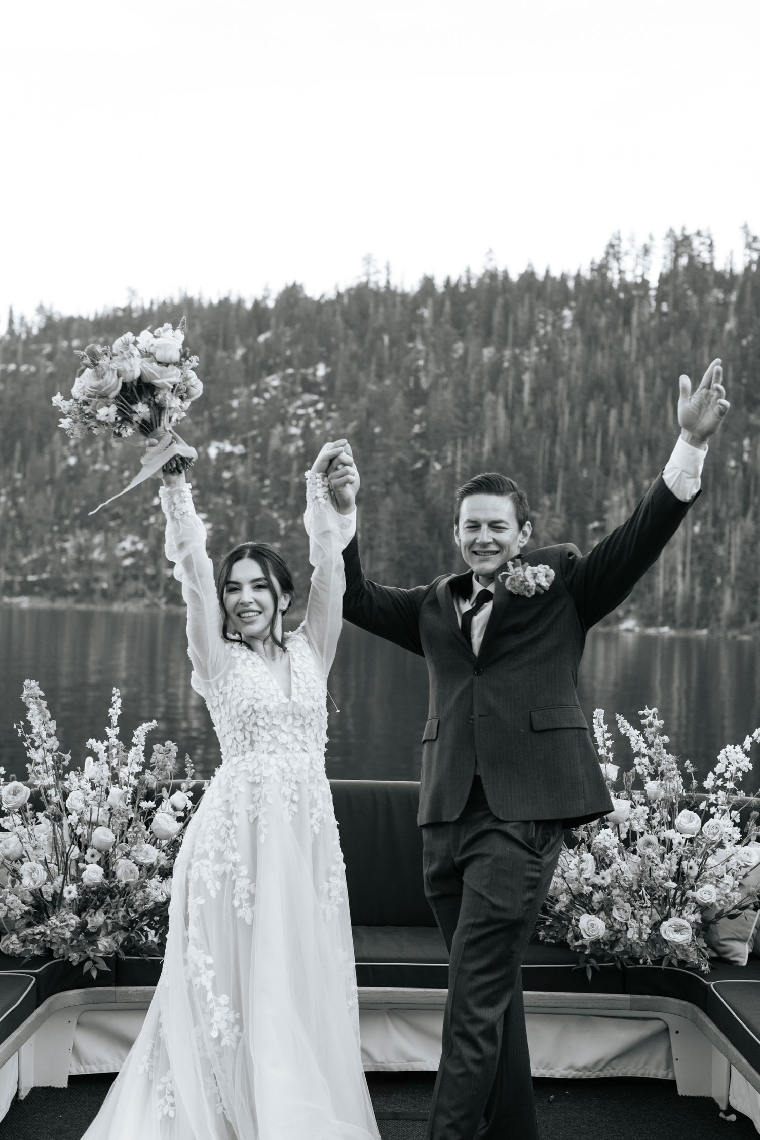 Wedding couple celebrating with raised arms against mountain lake view.
