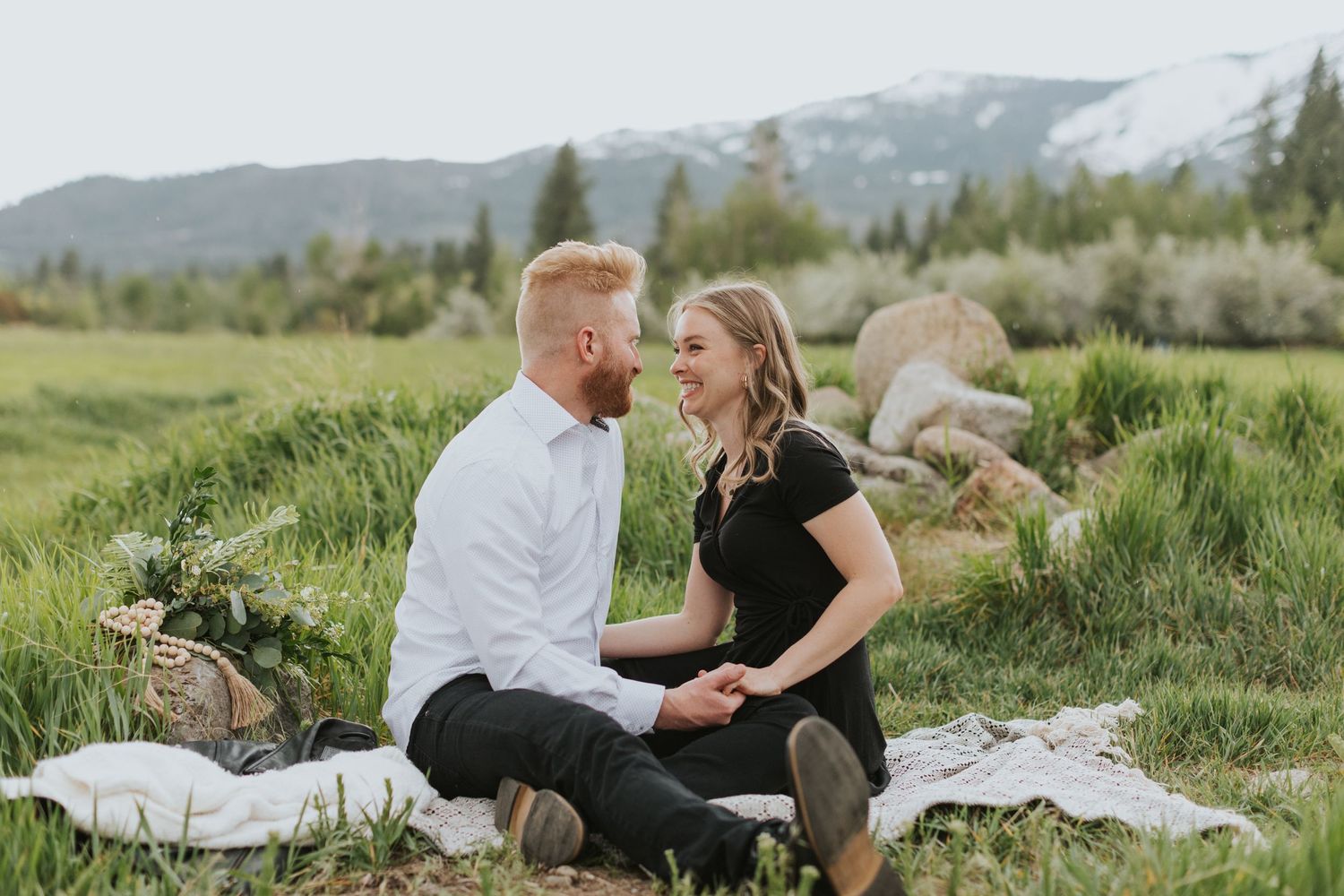 A couple sits together on a white blanket in a scenic mountain meadow during a romantic outdoor photoshoot.