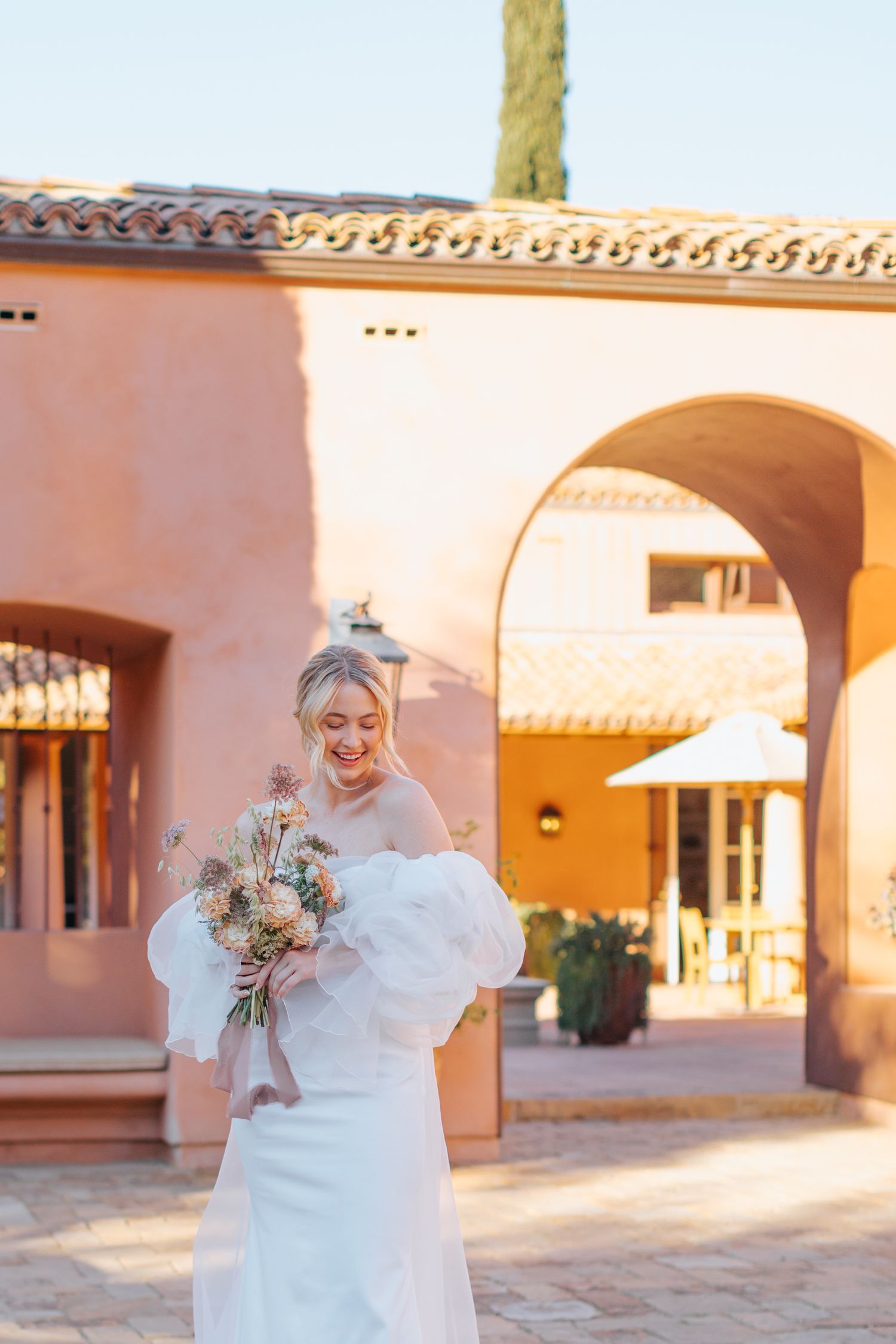 Person in white bridal gown holding bouquet in Spanish-style courtyard at sunset.