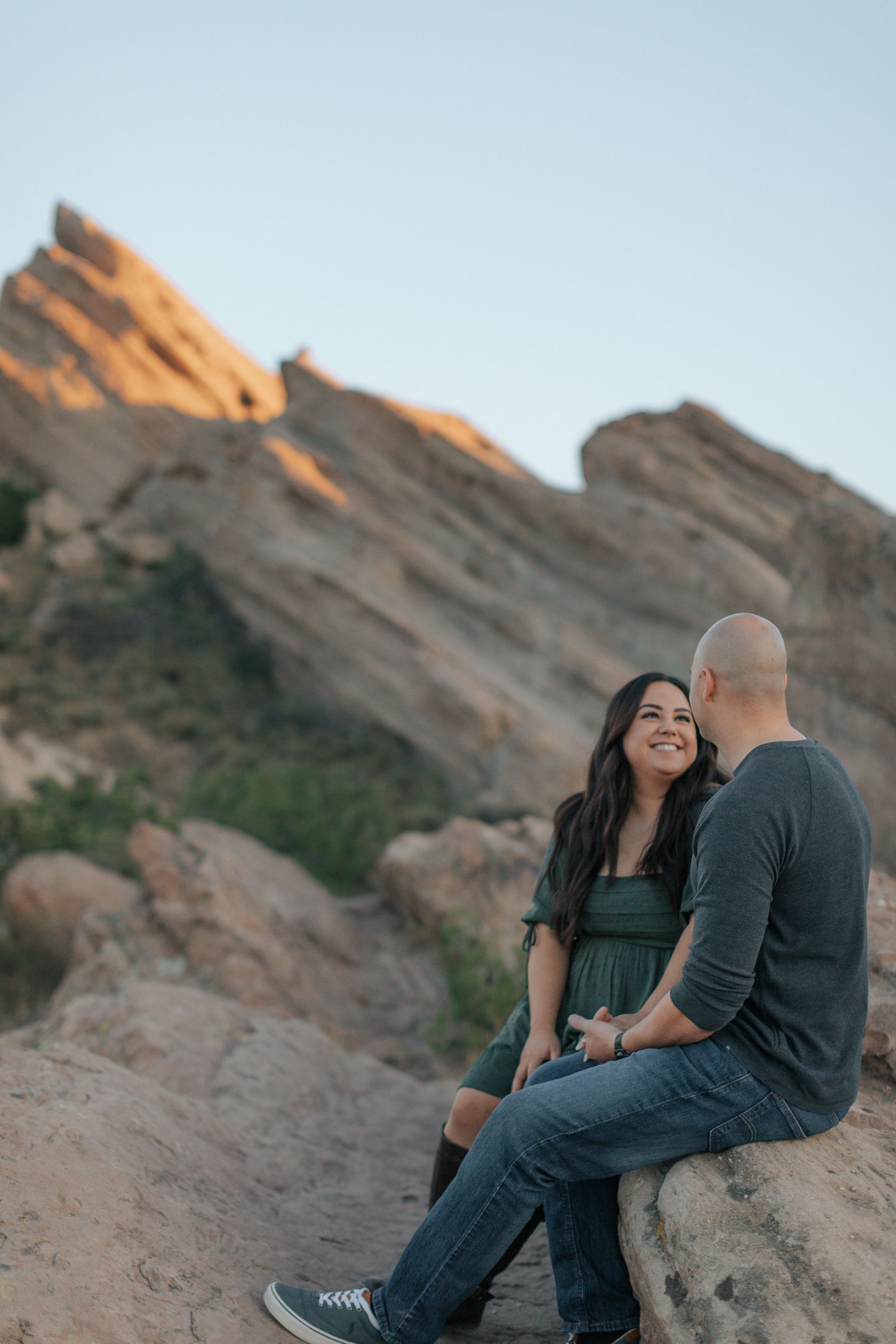Two people share a romantic moment while sitting on desert rocks during golden hour.