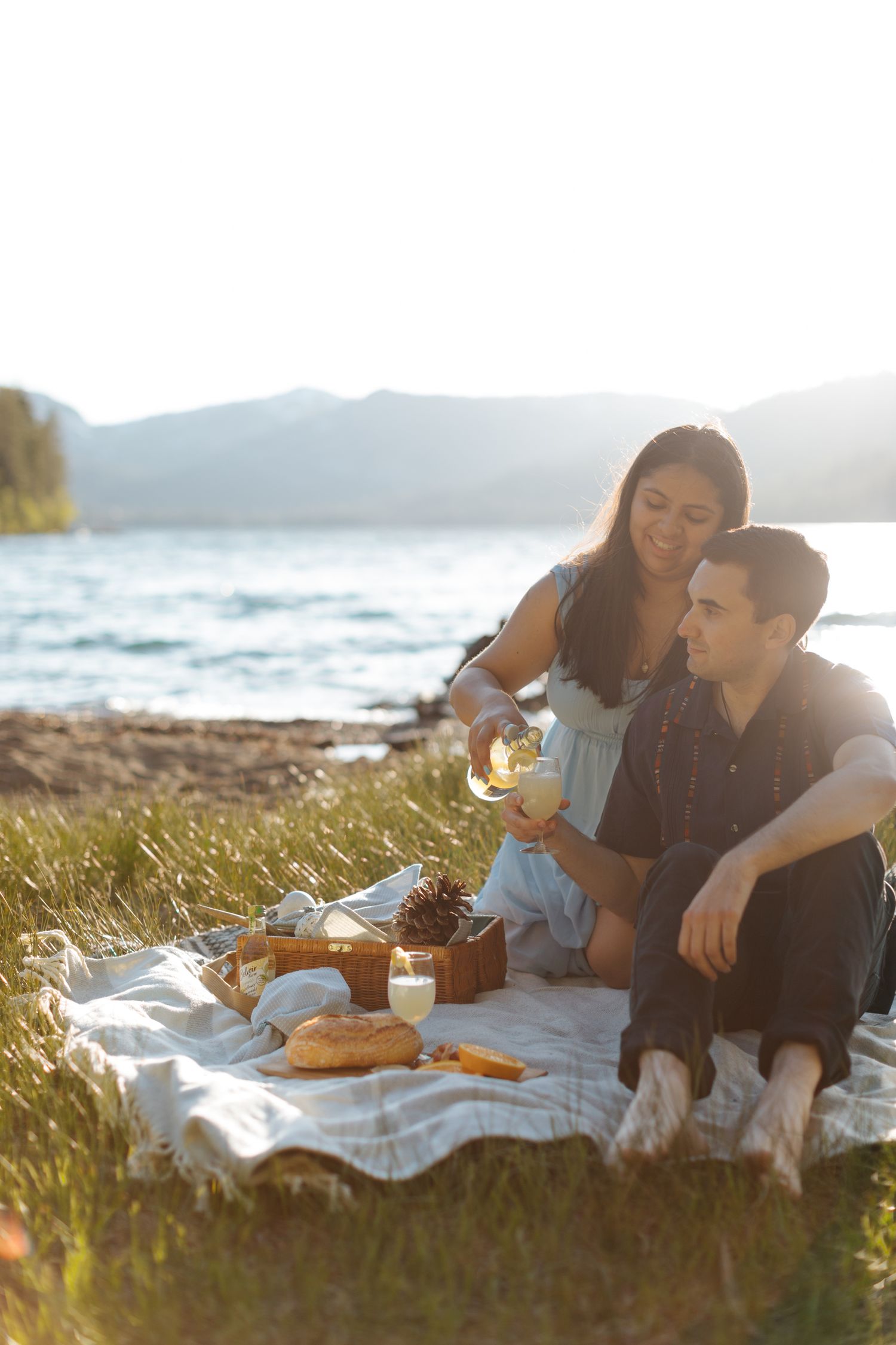 Couple having romantic picnic by ocean during golden hour with food spread on blanket.