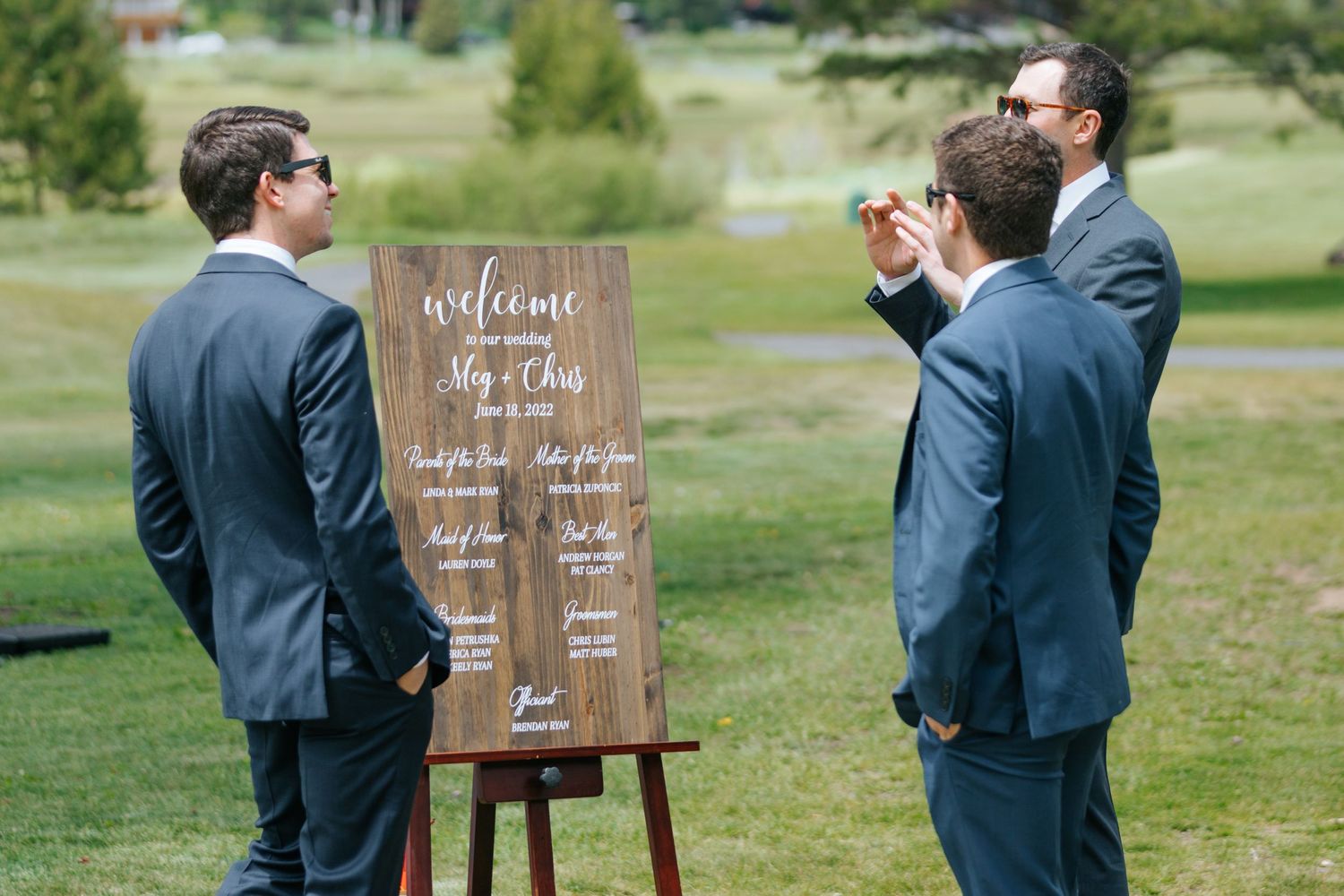 Groomsmen gather around wooden welcome sign at outdoor wedding venue.