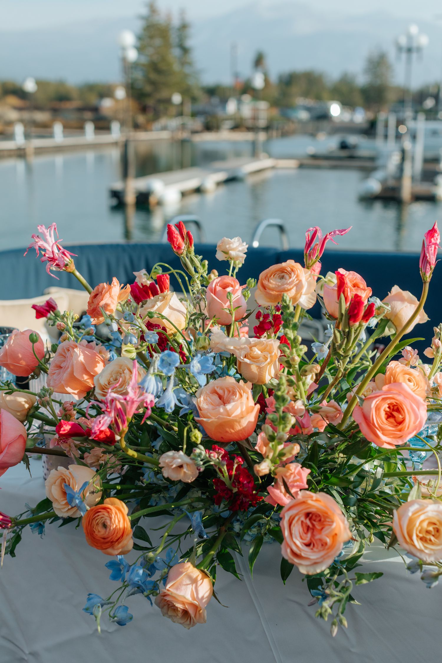 Vibrant coral and peach rose floral arrangement with blue accents against a marina backdrop.
