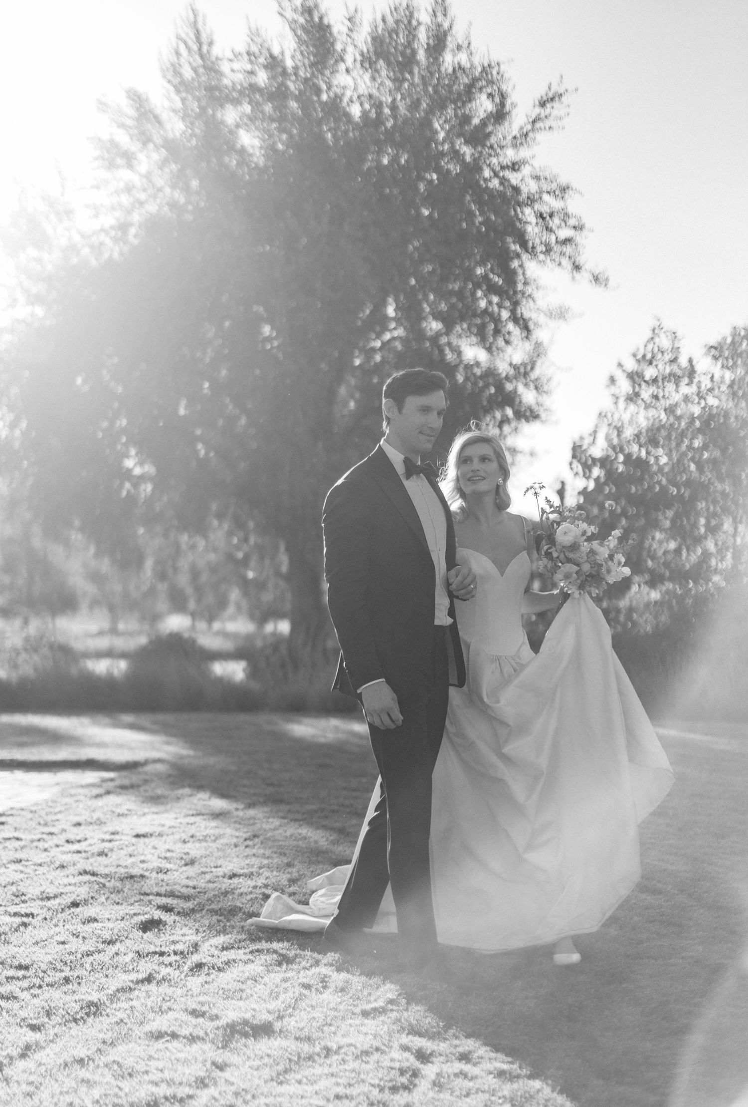 Black and white photo of a bride and groom walking outdoors on their wedding day with sunlight and trees in the background.