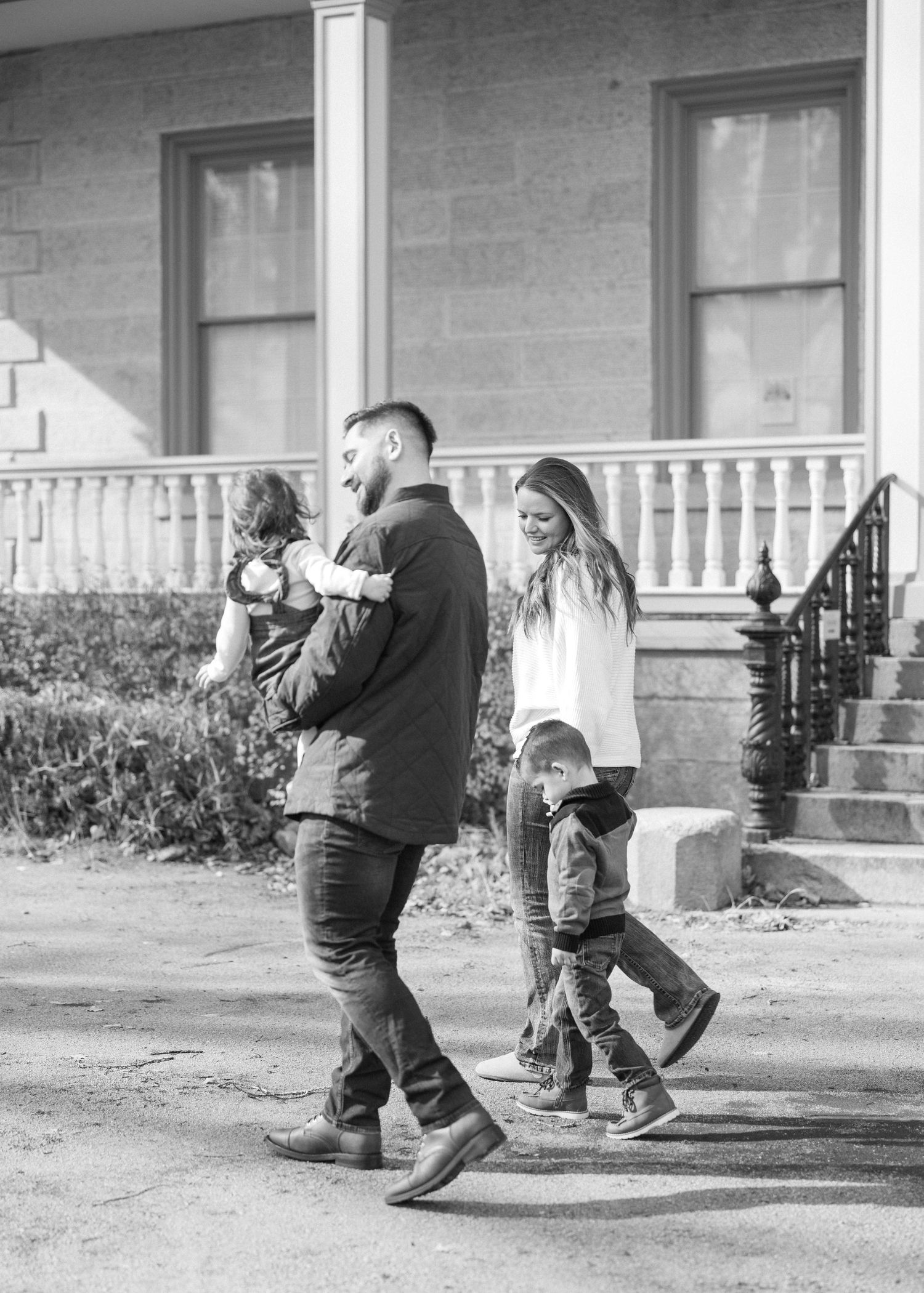 A family with two young children walks together outside a classic home in a black and white photo.