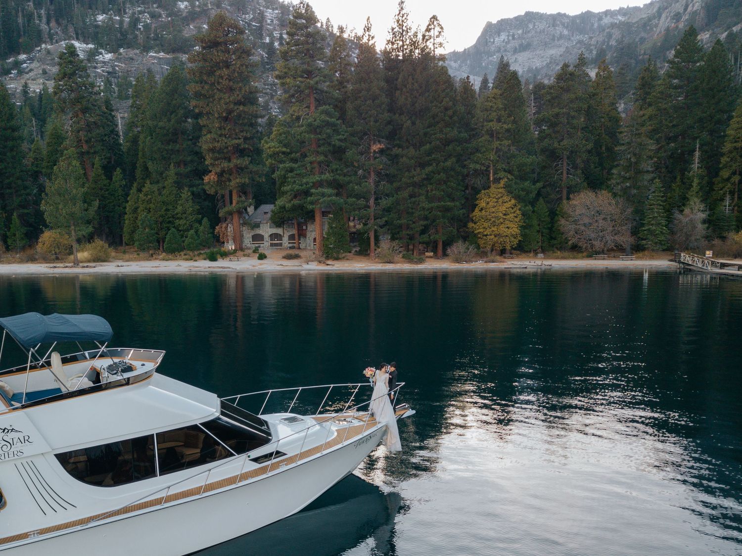 White boat cruising on a mountain lake surrounded by evergreen forest and snow-capped peaks.