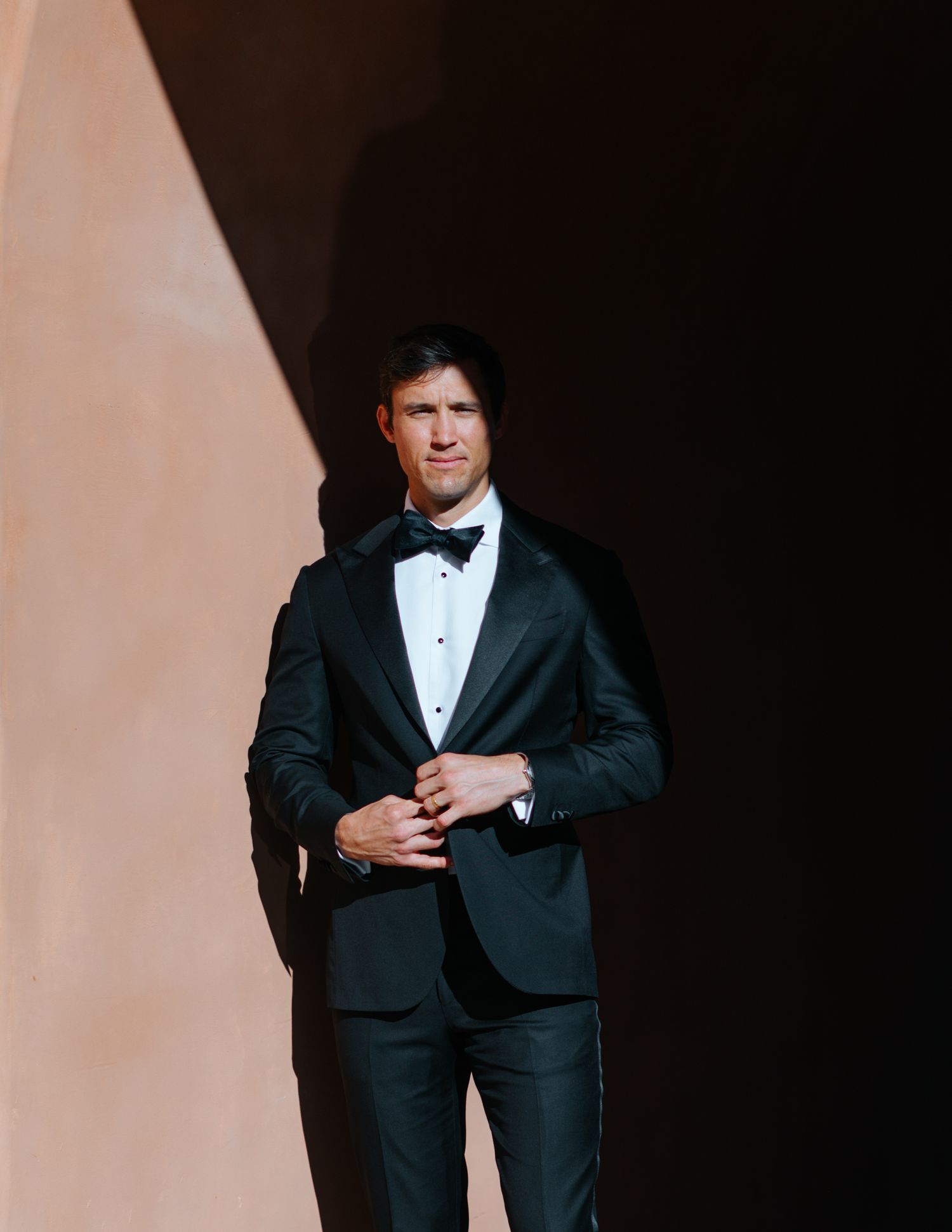 A man in a black tuxedo and bow tie poses against a light colored background in a series of dramatic shots.