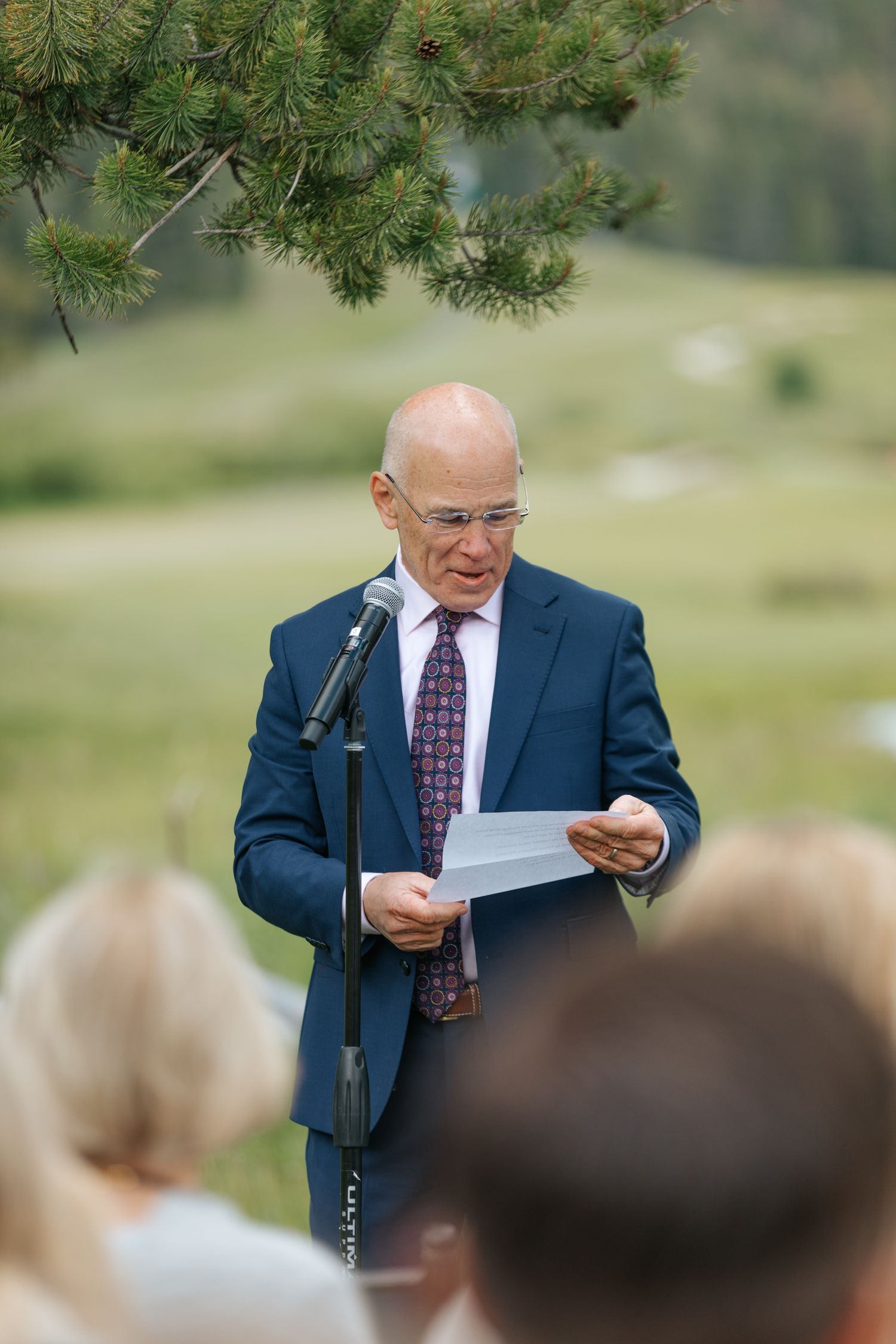 Speaker delivers remarks at outdoor wedding ceremony while holding papers.