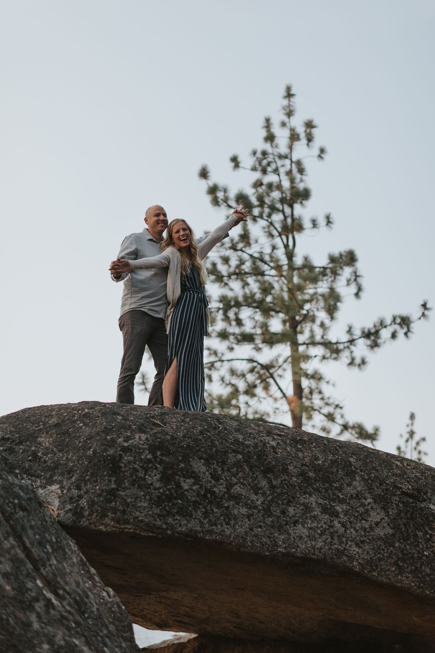 A couple stands on a large rock formation overlooking a scenic natural landscape with pine trees in the background.