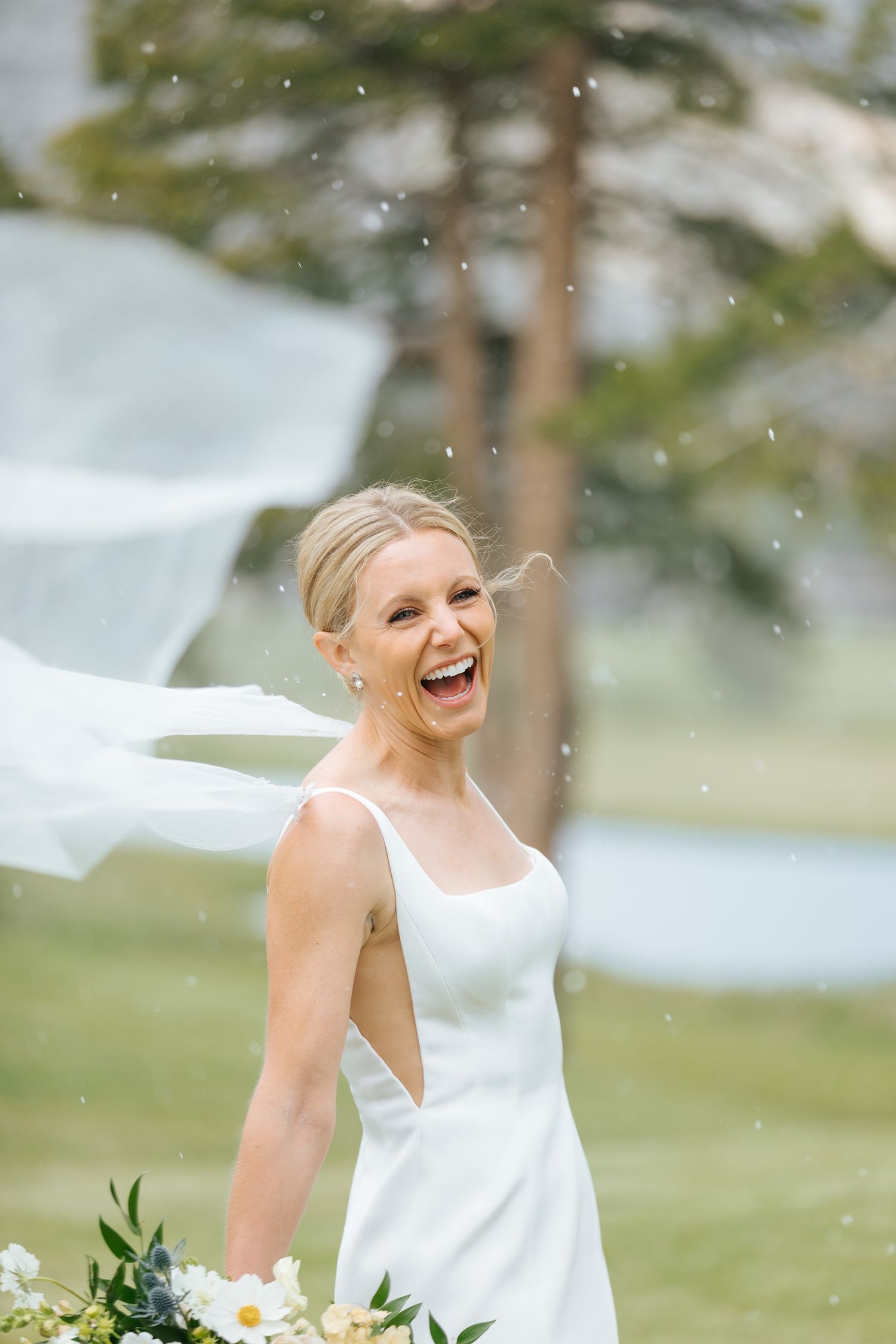 A bride in a sleek white dress laughs joyfully as her veil catches the wind on a sunny day.