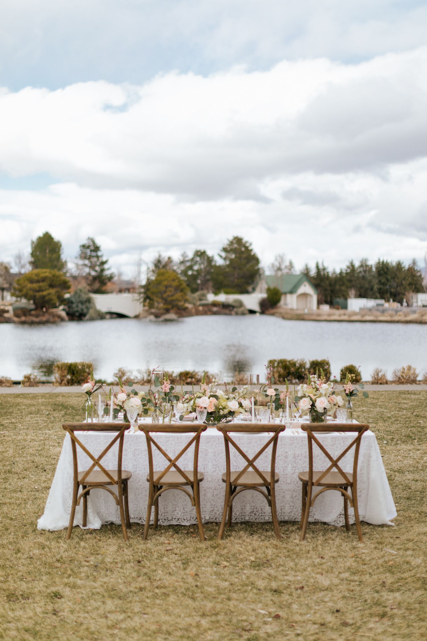 Rustic outdoor wedding reception table setup with cross-back chairs overlooking a lake.