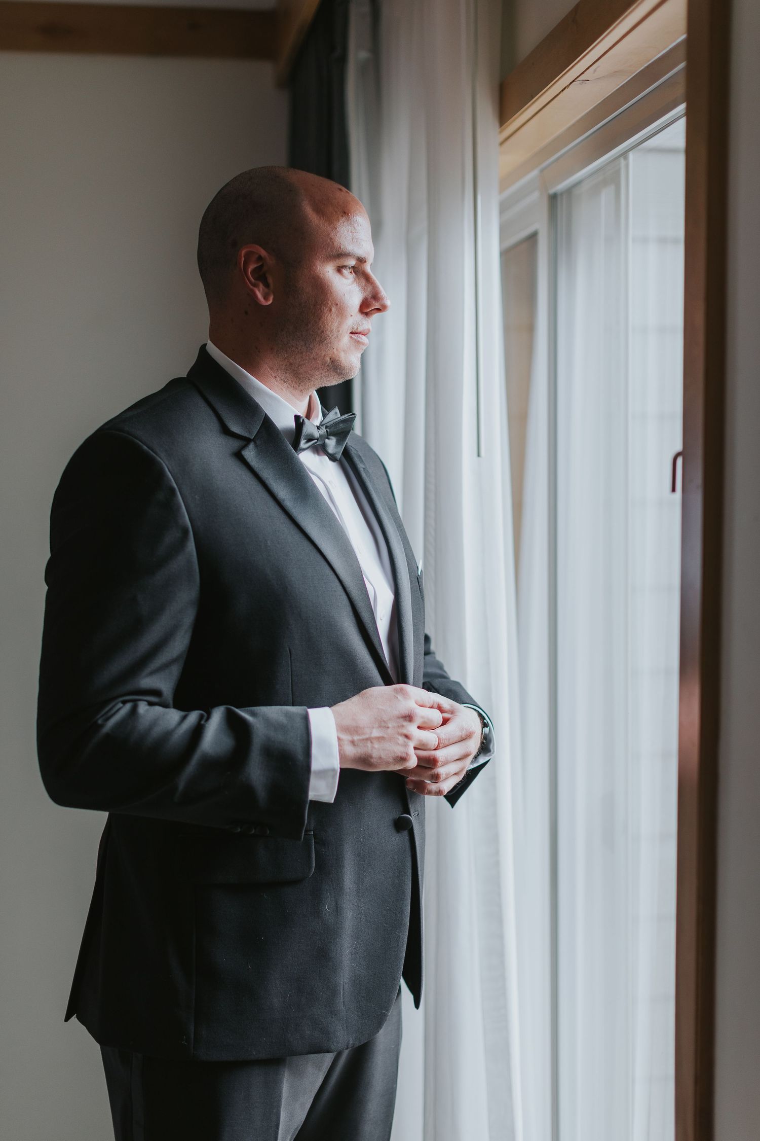 Groom in formal attire stands by window during wedding preparation.