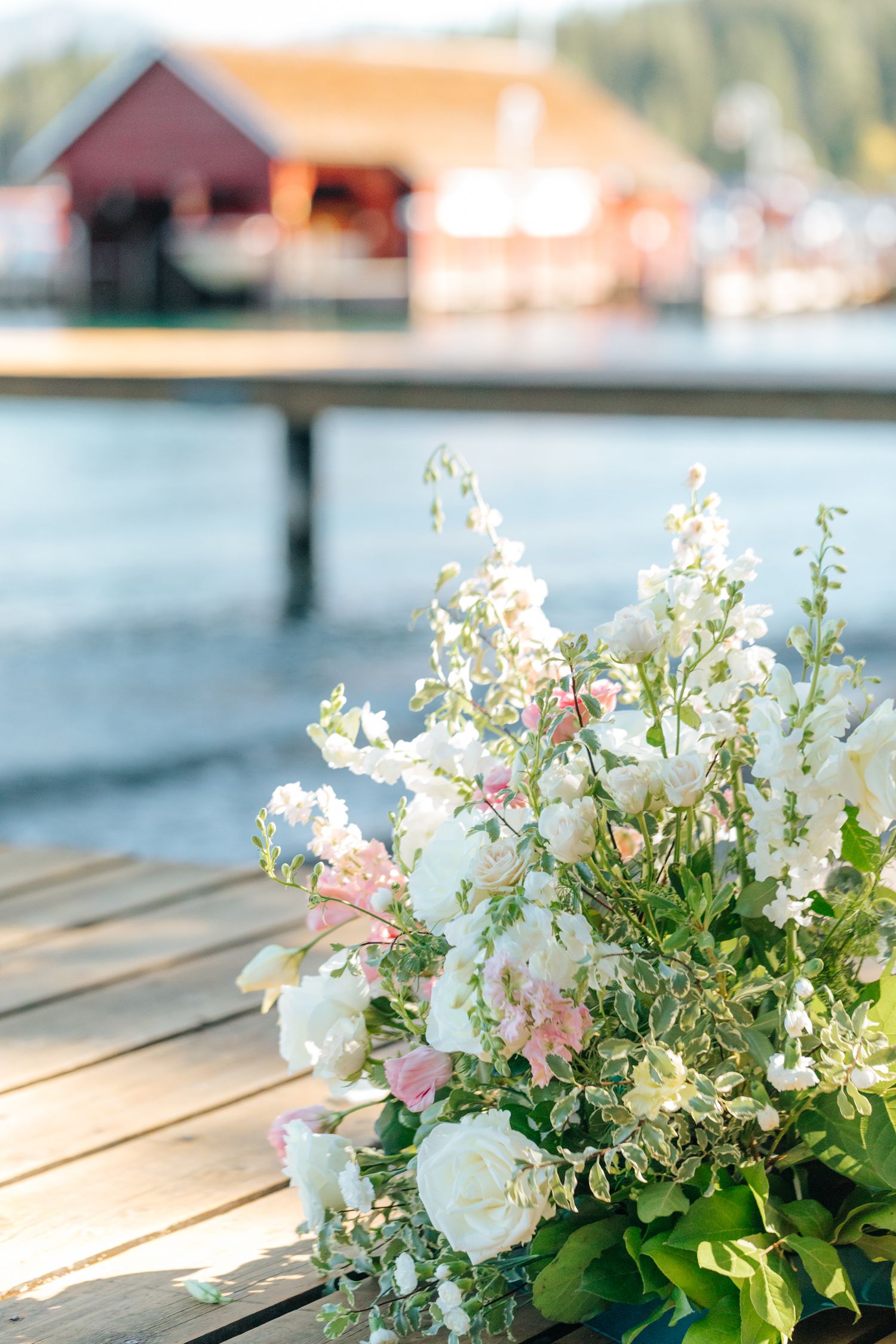 Delicate white and pink floral arrangements with greenery sit on a wooden deck overlooking a waterfront view.
