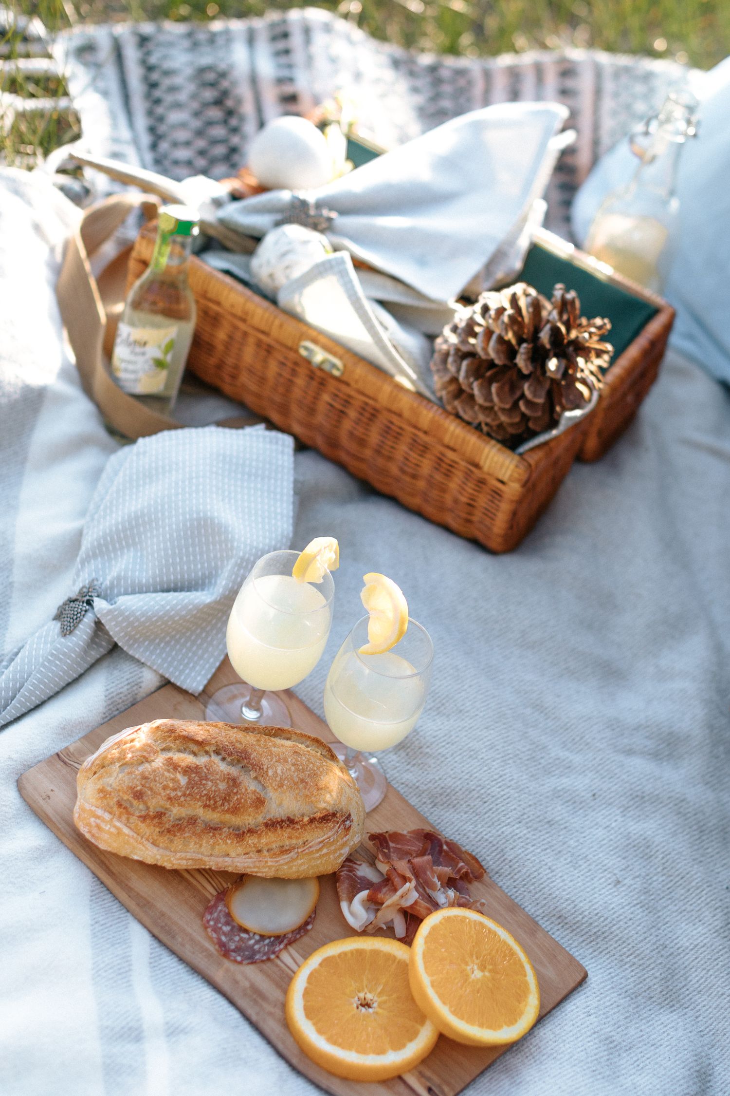 Picnic spread with fresh oranges, bread, and drinks on a gray blanket outdoors.
