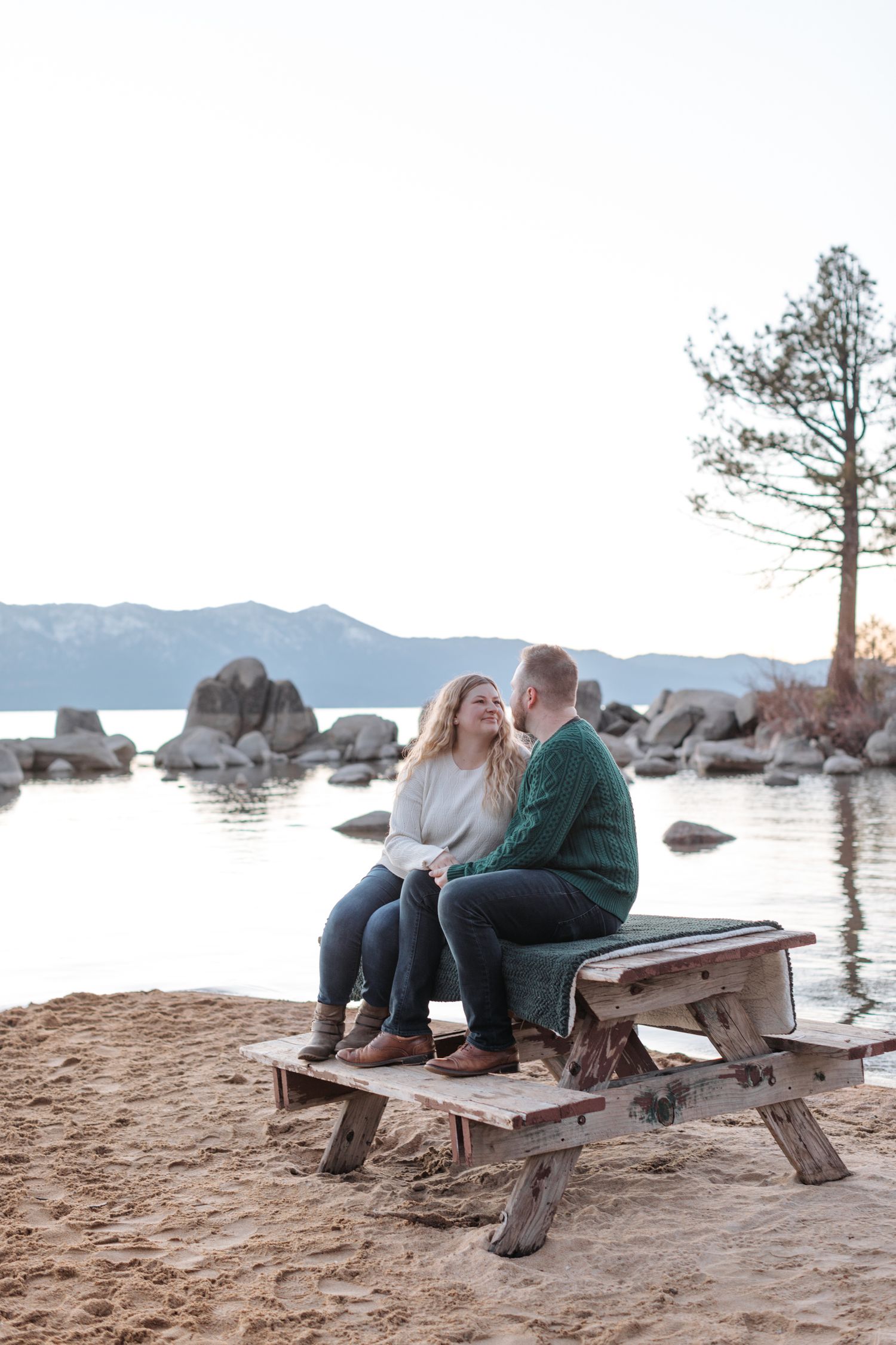Couple relaxing on a wooden bench at a scenic beach with mountain views and scattered rocks.
