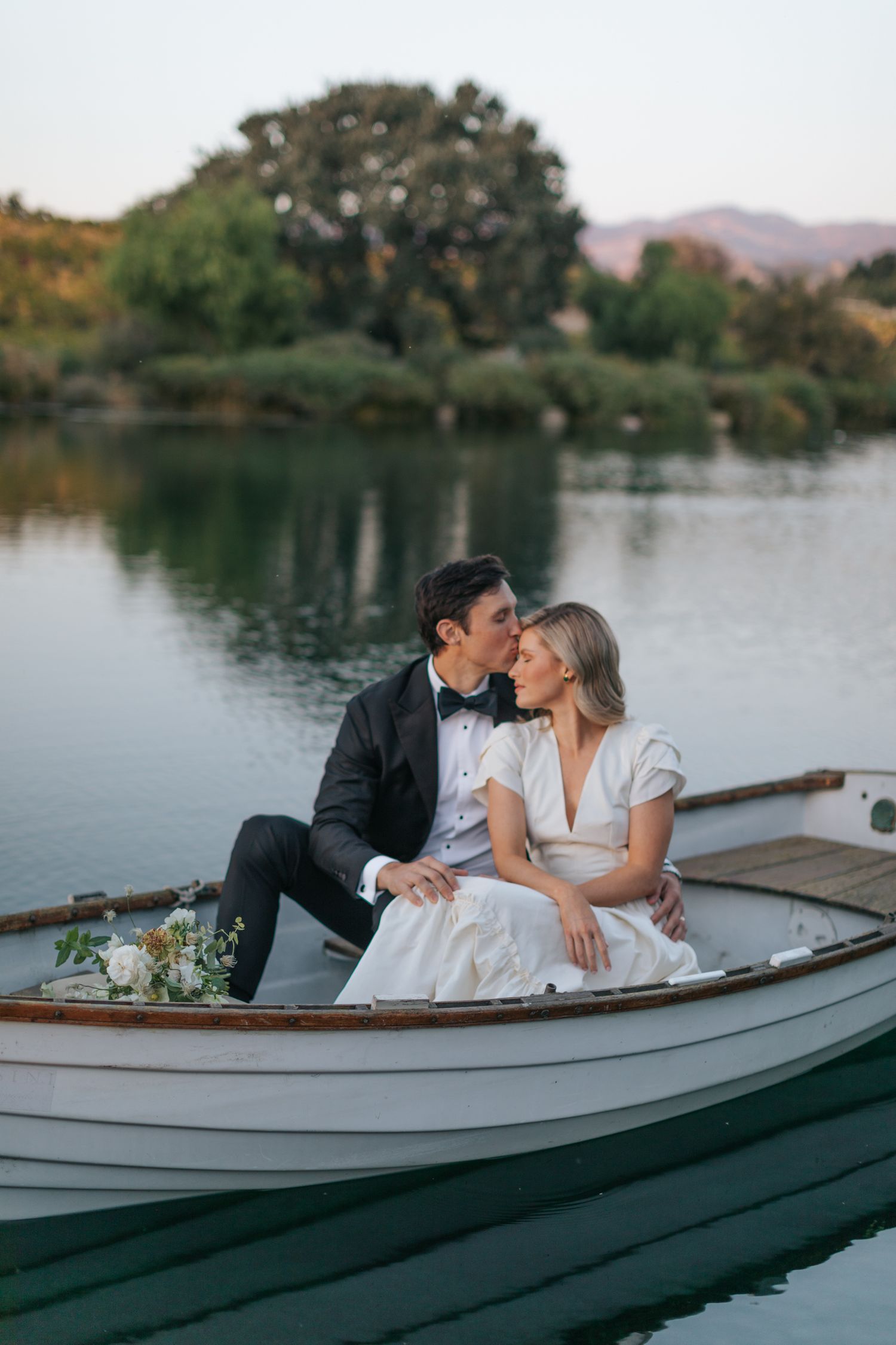 Romantic couple in wedding attire sitting in a rowboat on a serene lake.