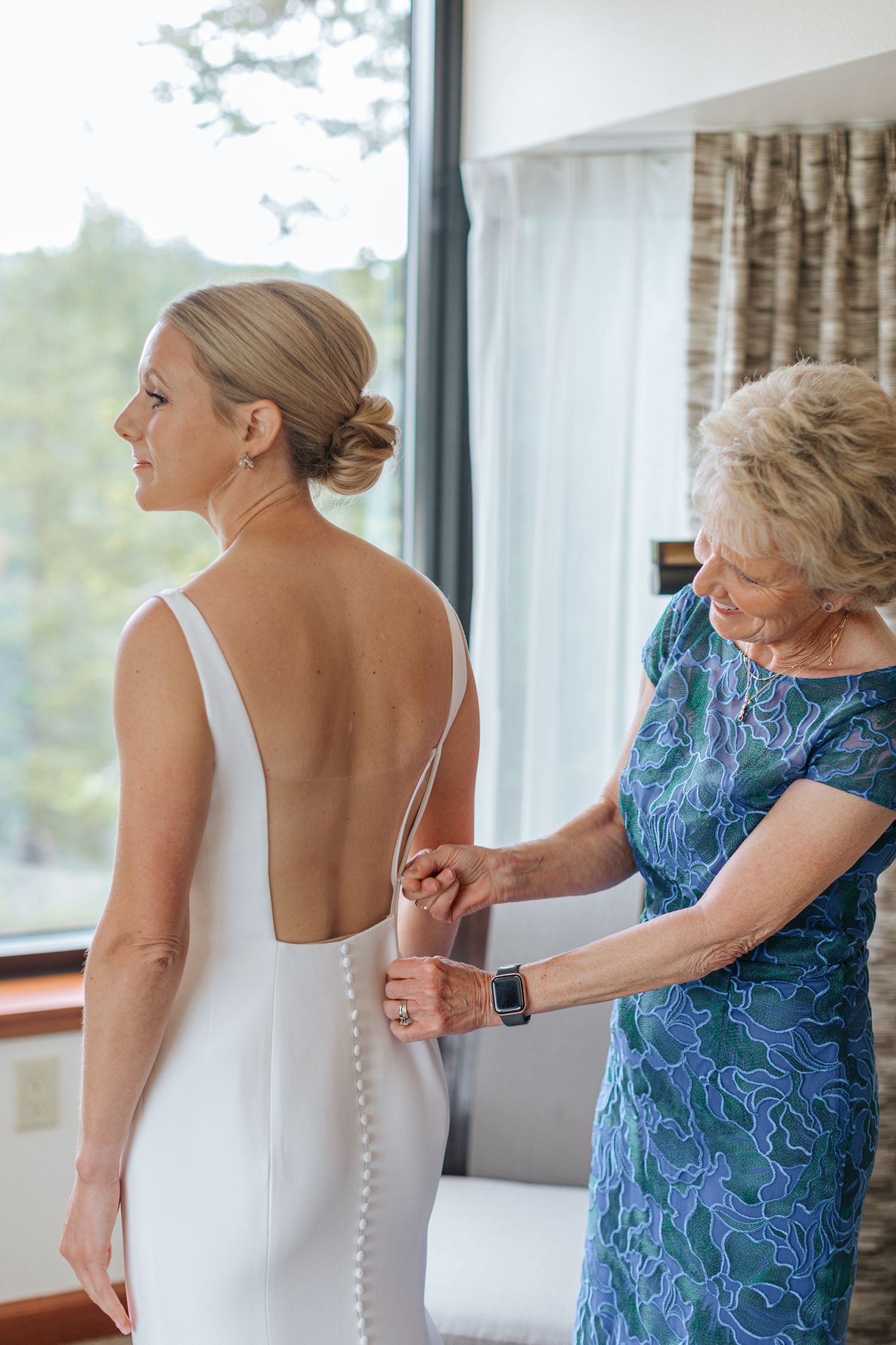 A woman in a white dress gets help with buttons while getting ready by a window.