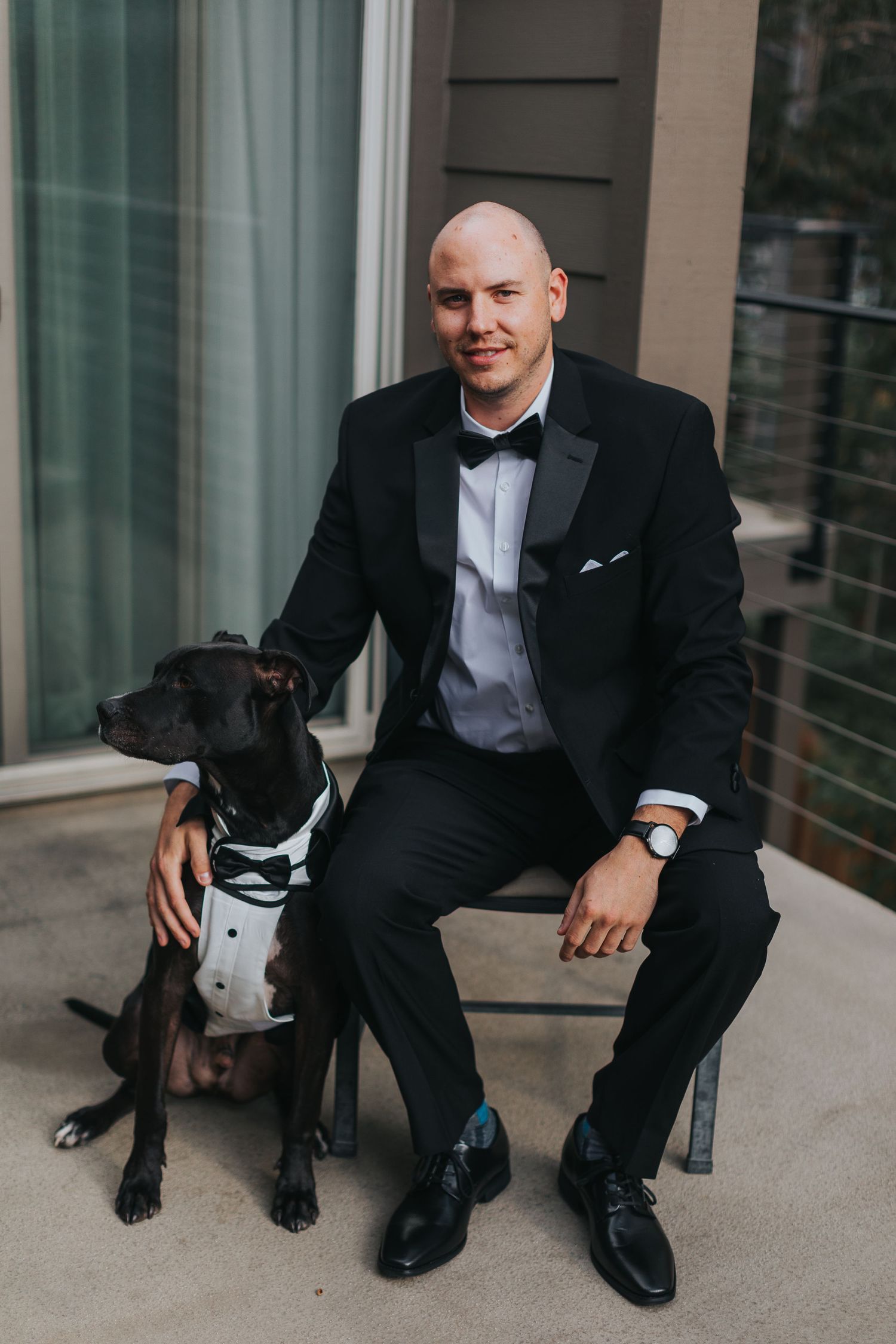 Person in tuxedo sitting with black dog on outdoor patio.