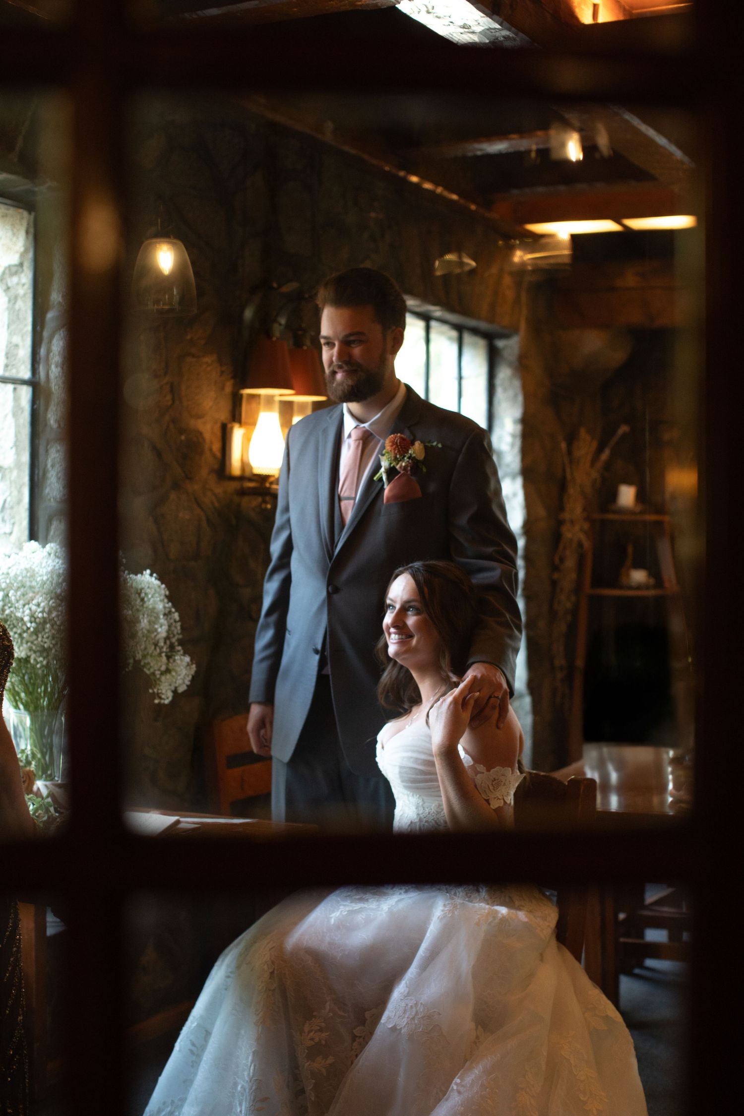 Bride and groom share a romantic moment inside a rustic stone venue, captured through a window frame.