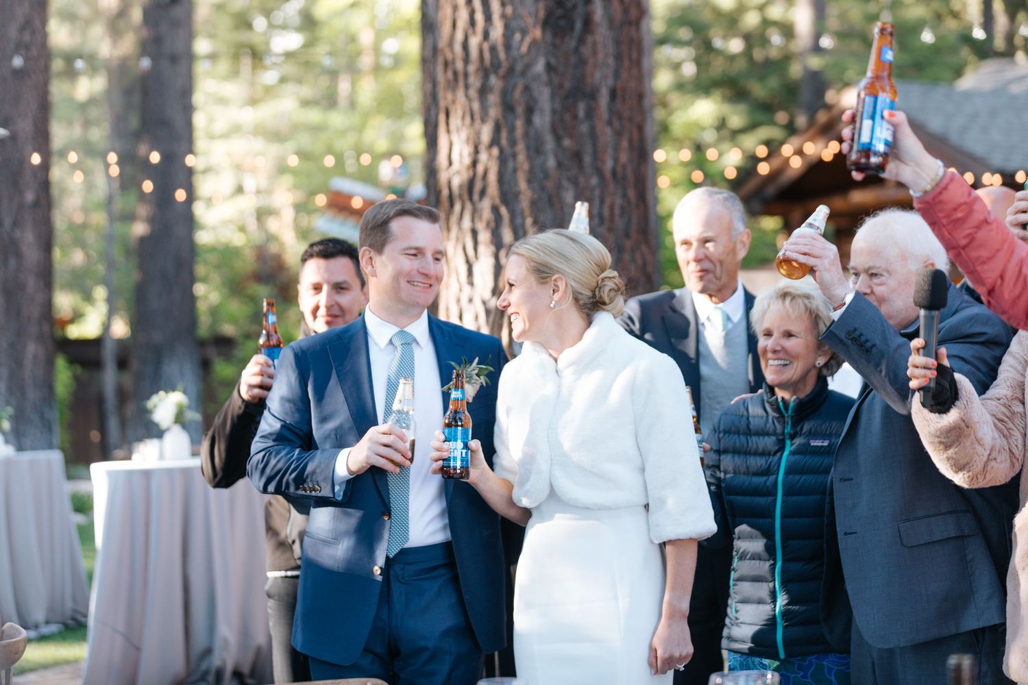 Bride and groom toast with guests outdoors at a rustic woodland wedding reception surrounded by tall pine trees.