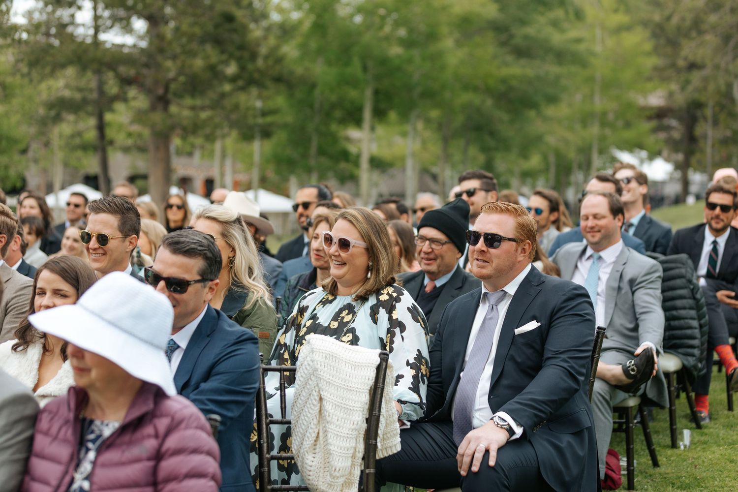 Wedding guests seated outdoors watch ceremony under trees on a sunny day.