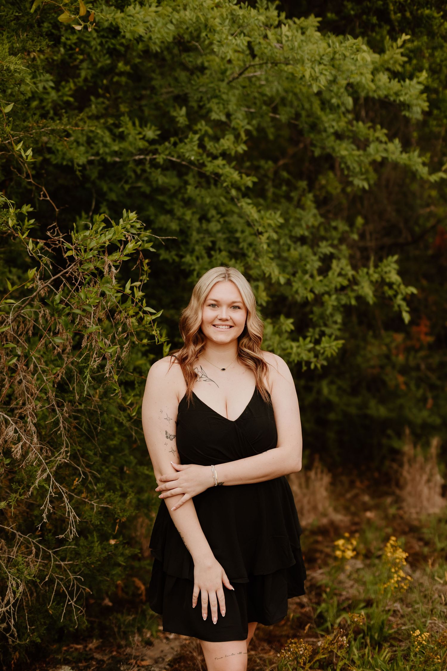 Young blonde woman in black dress smiling outdoors among lush green trees and foliage.