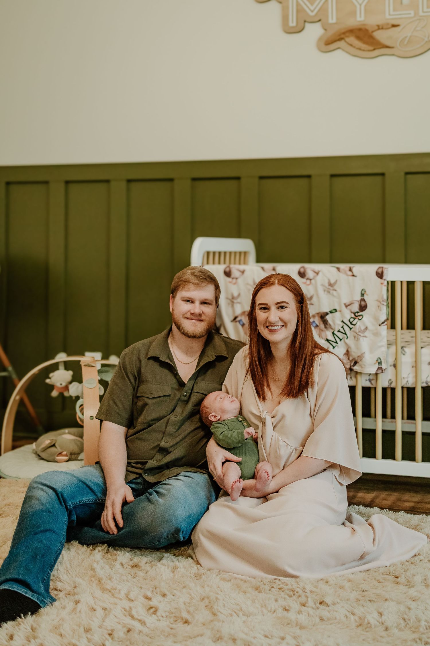 New parents sit on a fluffy rug holding their newborn baby boy Myles in a green nursery.