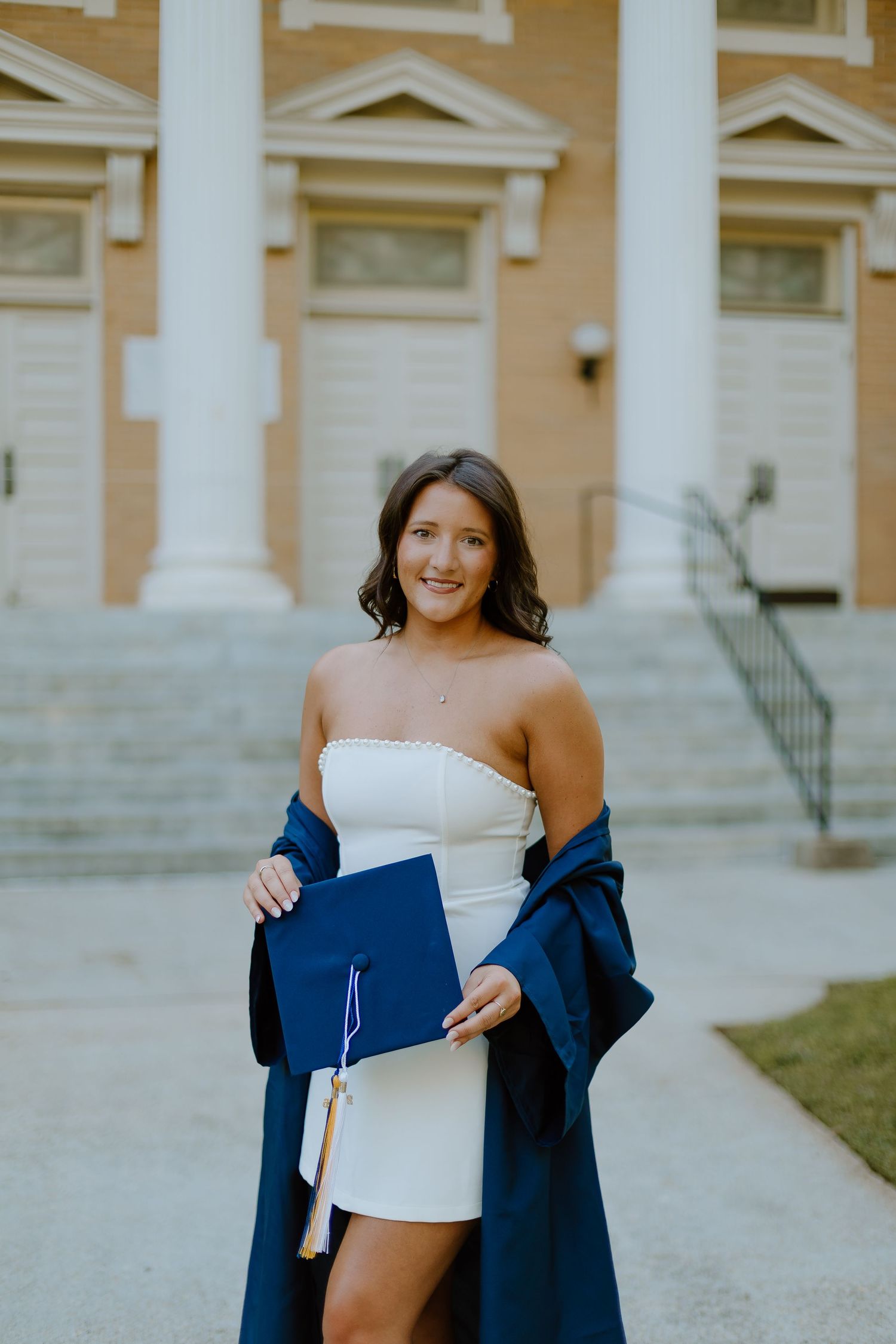 Smiling graduate in white dress holding navy cap and gown outside brick university building.