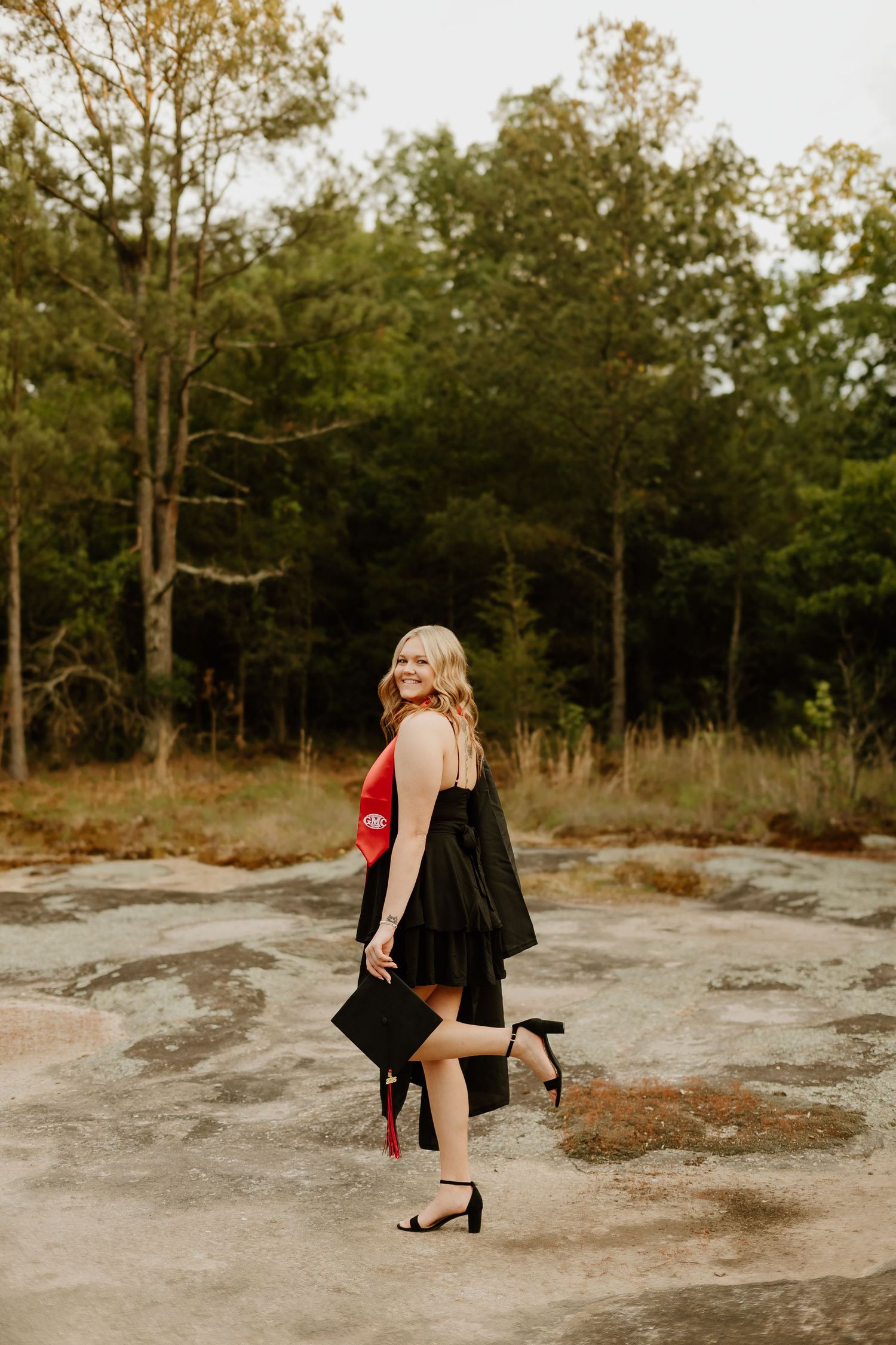 Smiling blonde graduate in black dress holds cap and red gown outdoors in a wooded field during golden hour.