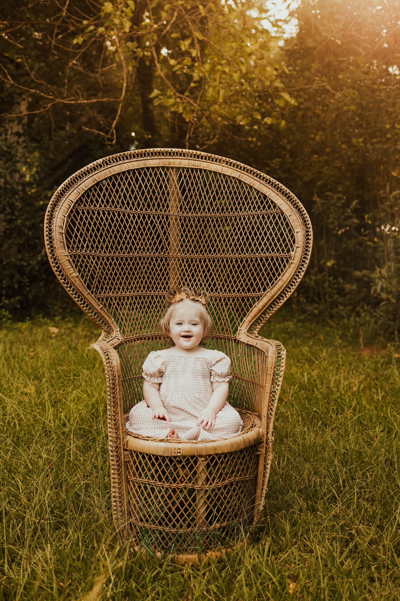 Happy baby girl in a white dress laughing while sitting in a large wicker peacock chair on green grass.
