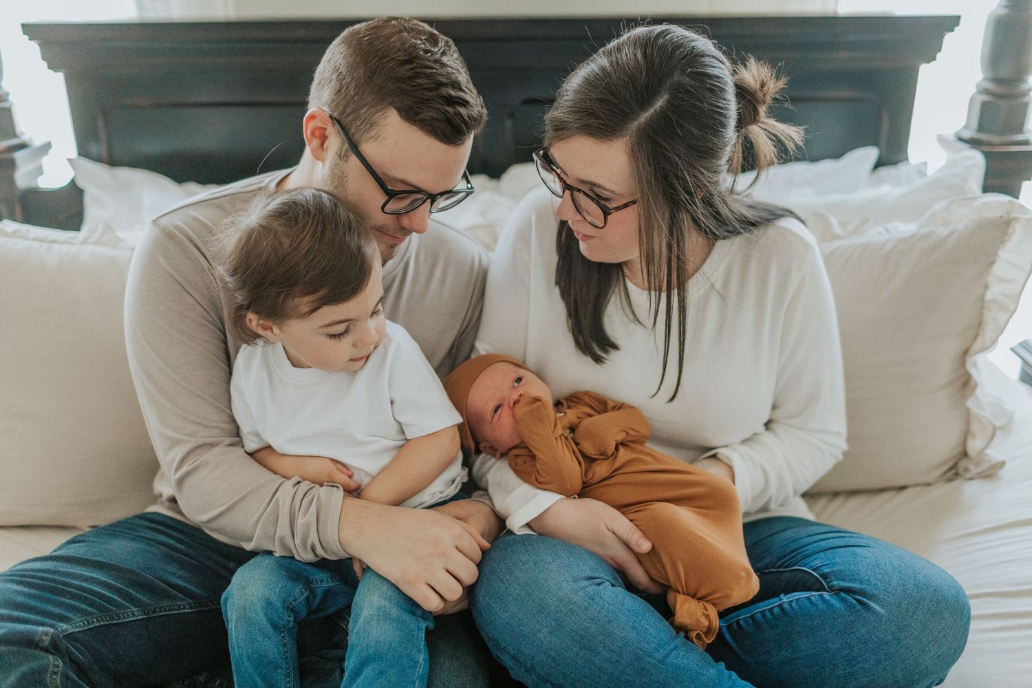 Happy family of four sits on bed as parents and toddler lovingly look at swaddled newborn.
