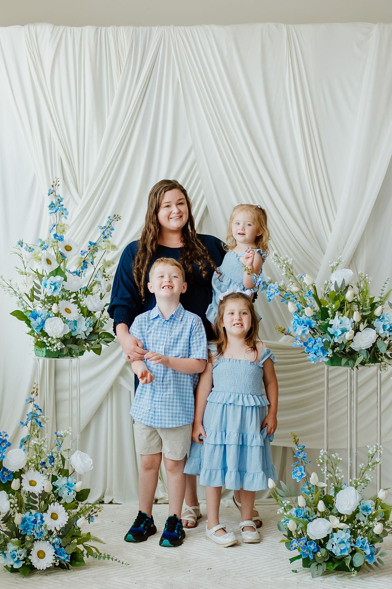 A woman poses with three young children in coordinating blue outfits before a white draped backdrop with blue and white floral arrangements.