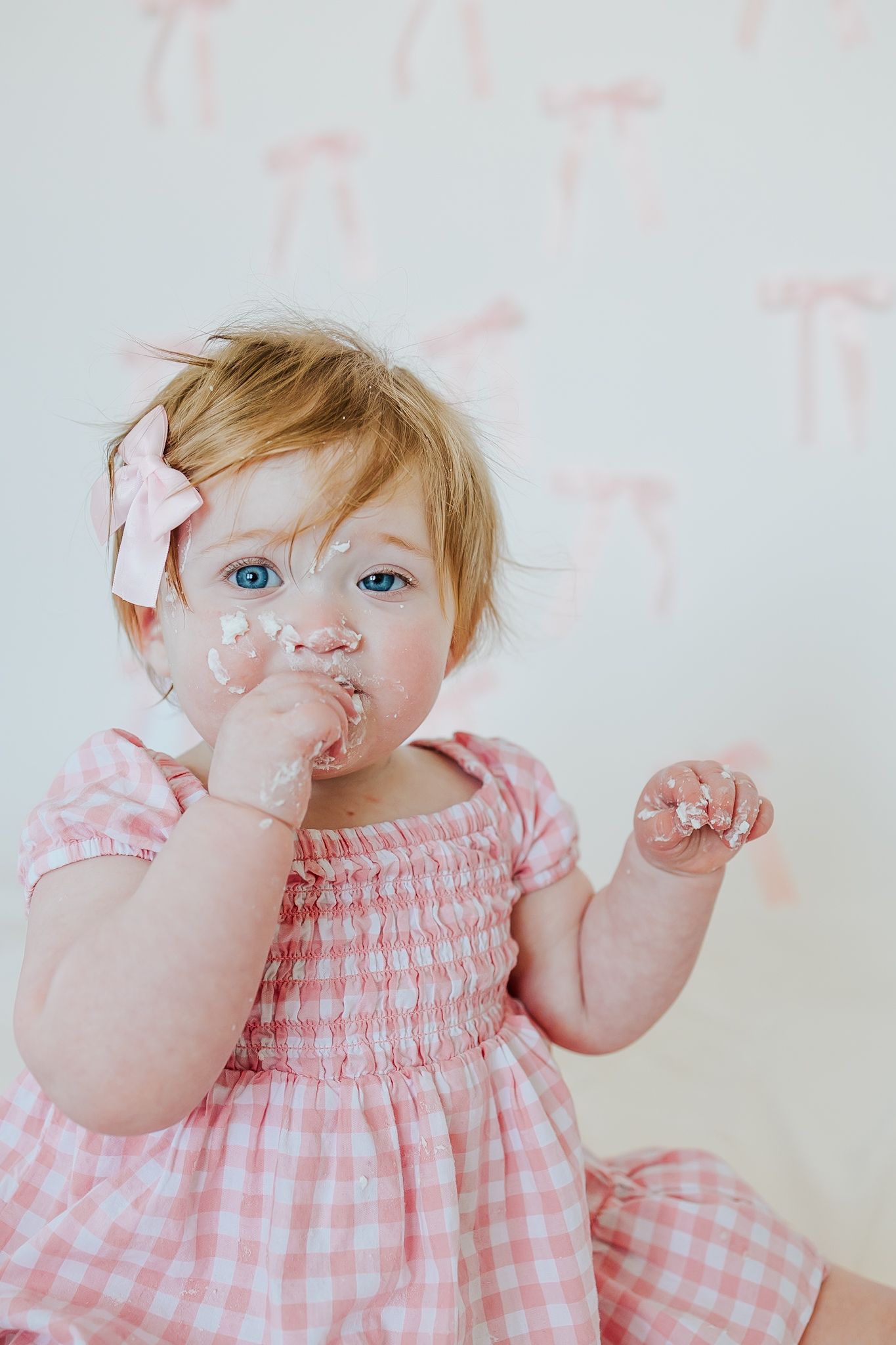 Adorable blue-eyed baby girl in pink gingham dress enjoys smash cake with frosting on her face.