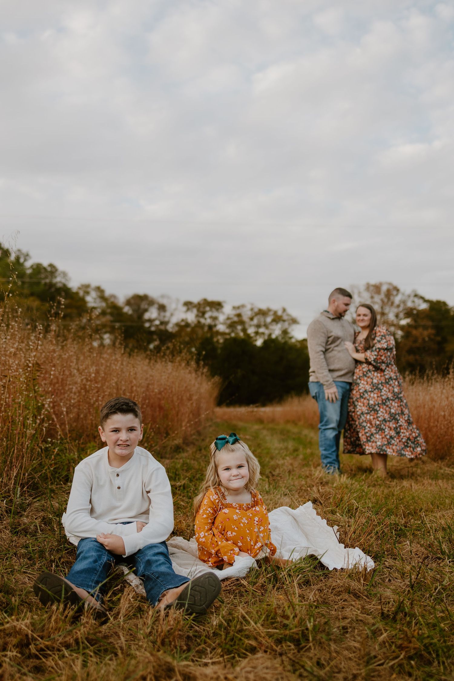 Two siblings sitting in grass while parents embrace in autumn field background.